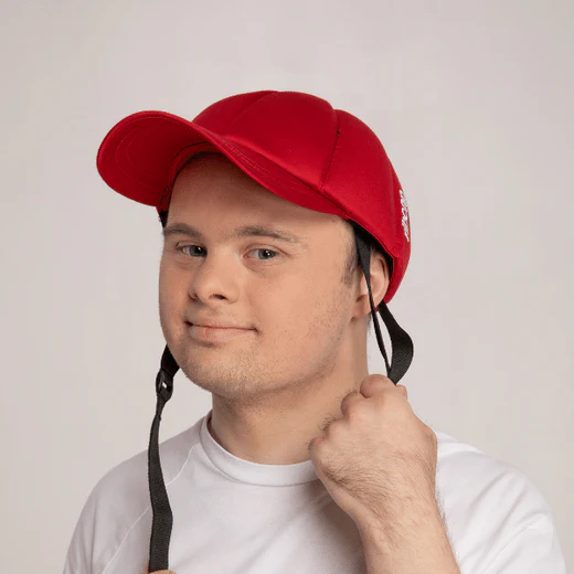 A young man wearing a red cap and a white t-shirt is smiling slightly while holding one of the black straps of his cap in his hand. The background is plain and light-colored.
