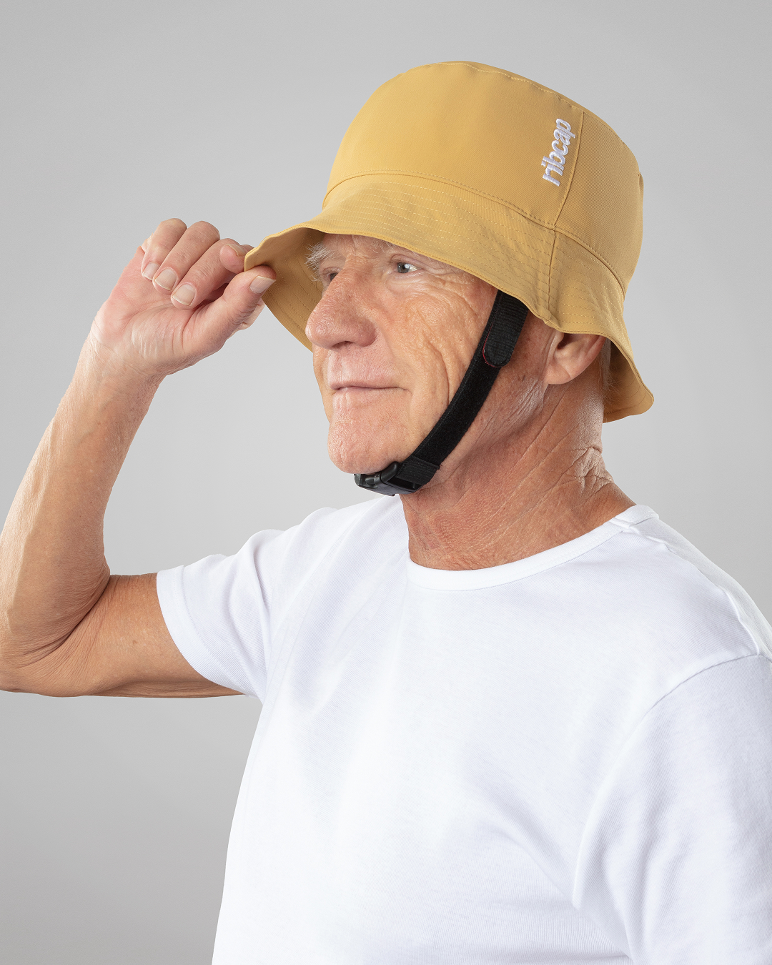 An older man in a white t-shirt smiles and adjusts a yellow bucket hat with a chin strap. The background is plain gray.