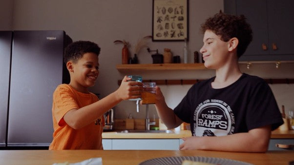 Two boys sit at a kitchen table, smiling and clinking their glasses together in a toast. The kitchen background includes shelves, plants, and a refrigerator.