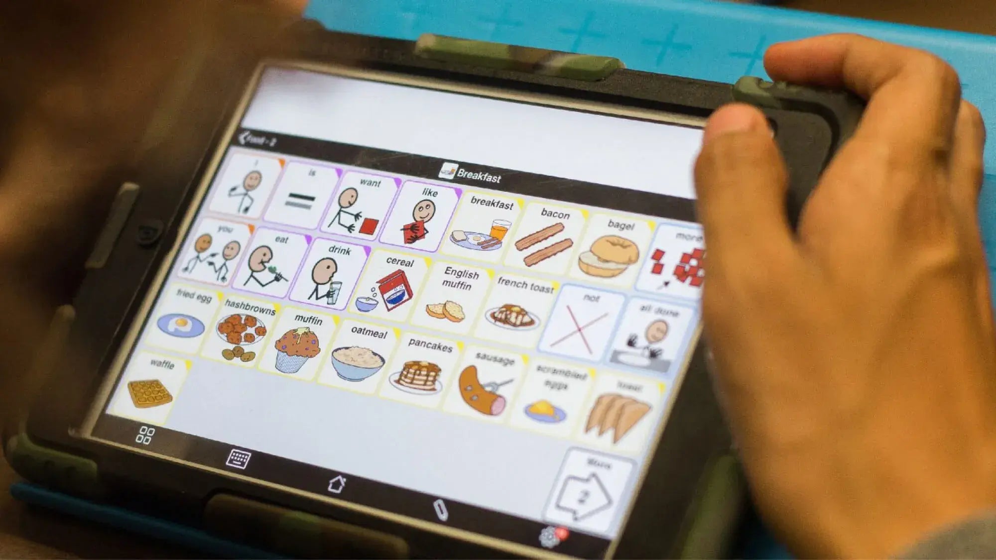 A person uses a tablet displaying a communication app with illustrated icons and words for food items like breakfast, oatmeal, pancakes, bacon, and eggs.