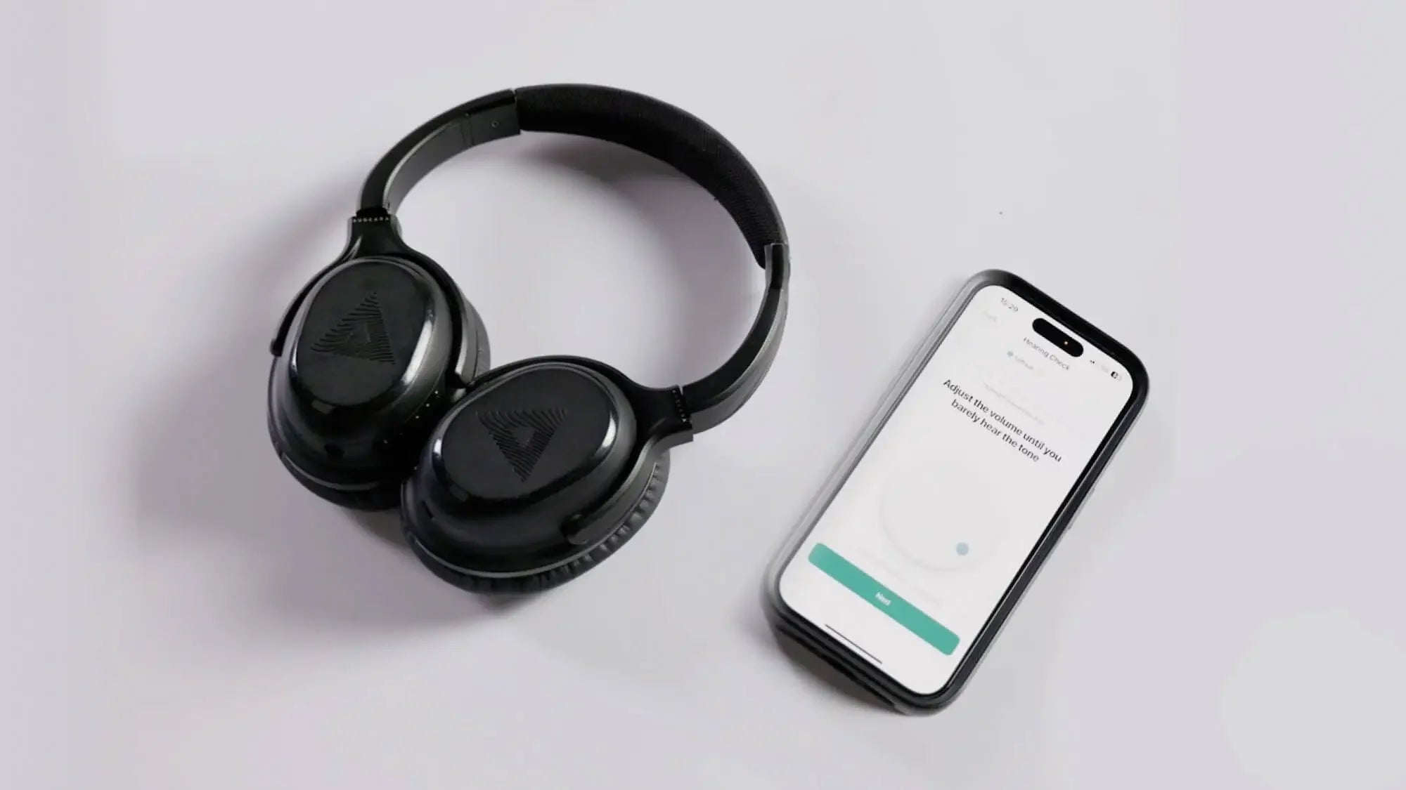 Black over-ear wireless headphones are placed next to a smartphone displaying a setup or pairing screen on a white surface.