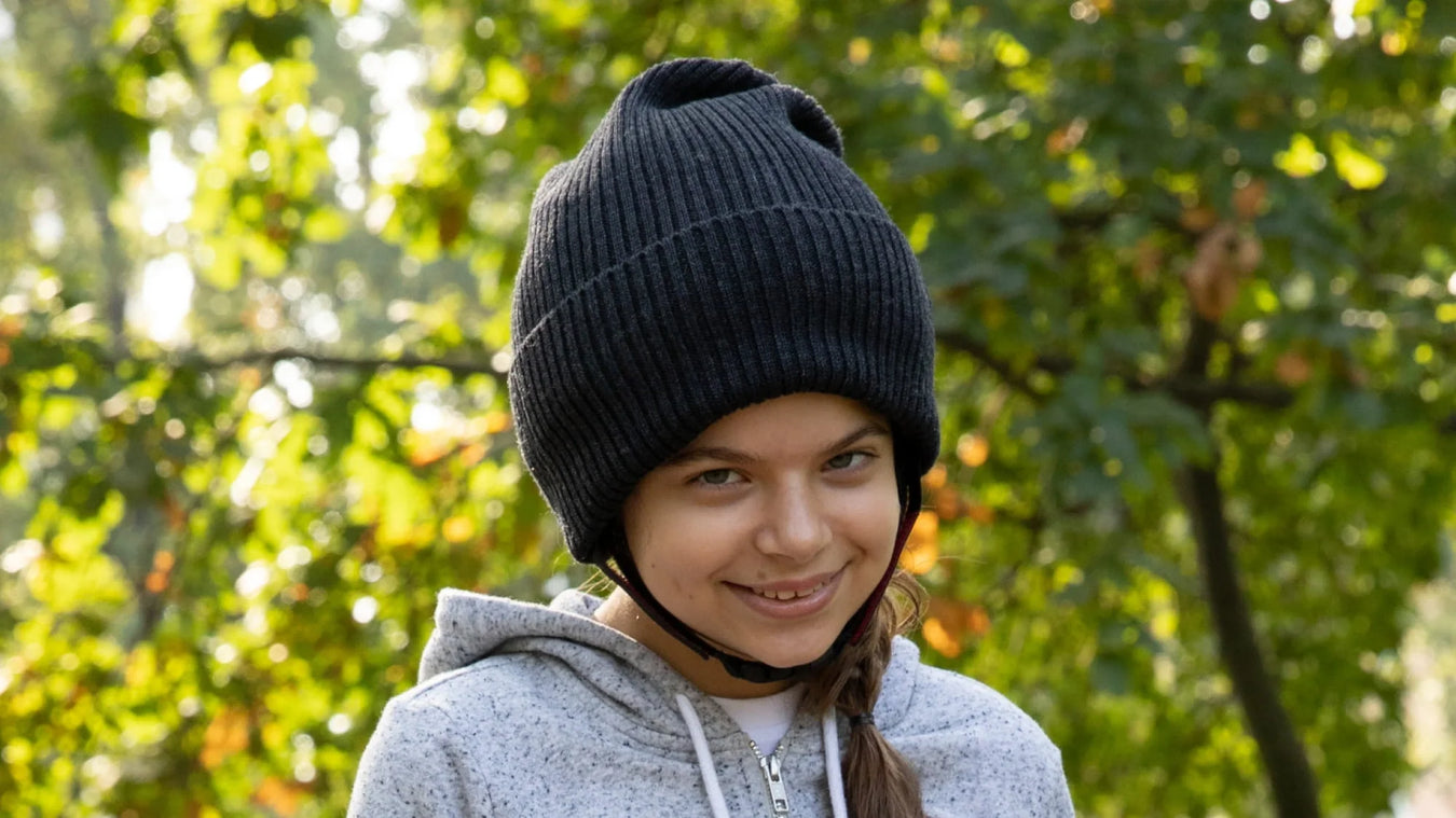 A young person with a long brown braid and a gray hoodie smiles while wearing a large black beanie hat outdoors, with green trees and sunlight in the background.