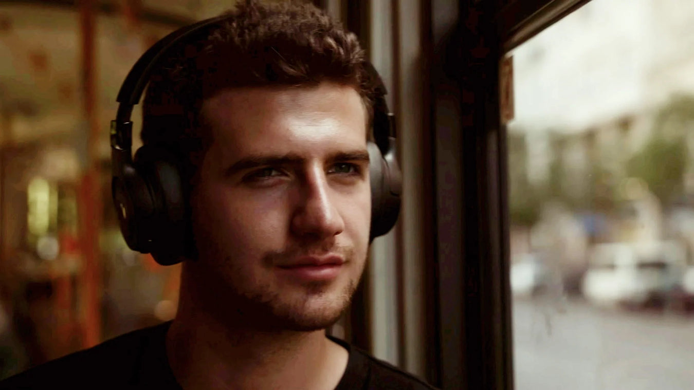 A young man with short dark hair and headphones gazes out of a window with a slight smile, sunlight illuminating his face. The background is softly blurred, suggesting he is on public transport.