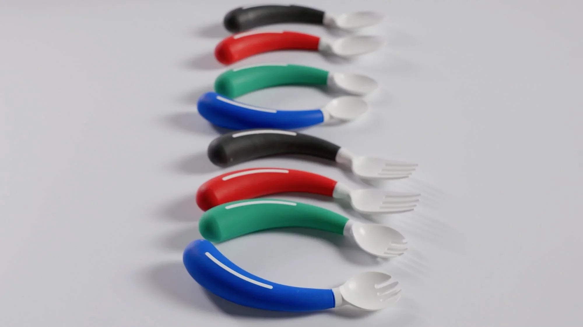 Six pairs of adaptive utensils, each with a curved handle and white plastic head, are arranged in rows. The handles are colored black, red, green, and blue, alternating in order.