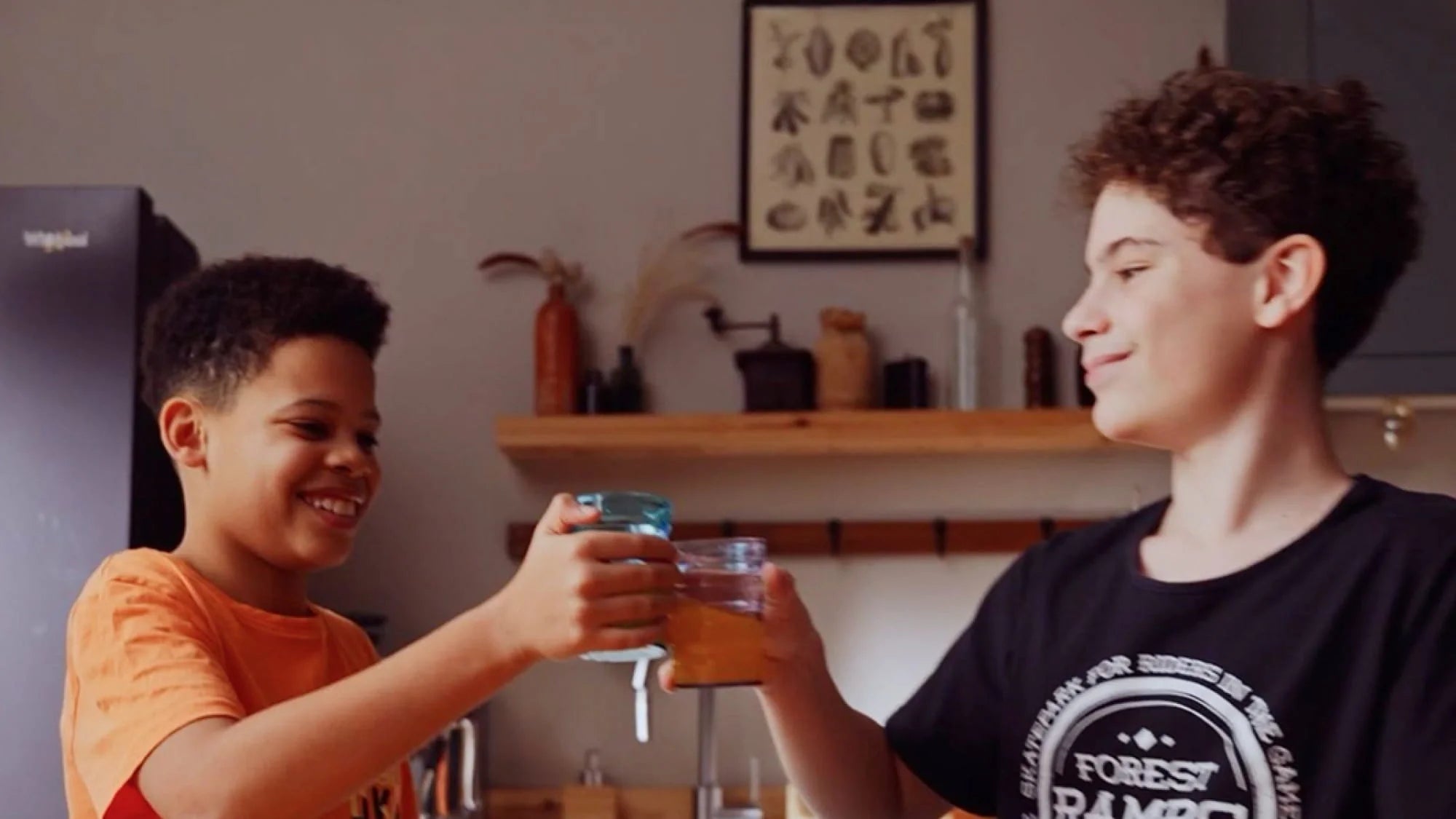Two boys smile and clink glasses together in a kitchen, appearing happy. Shelves, jars, and framed art are visible in the background.