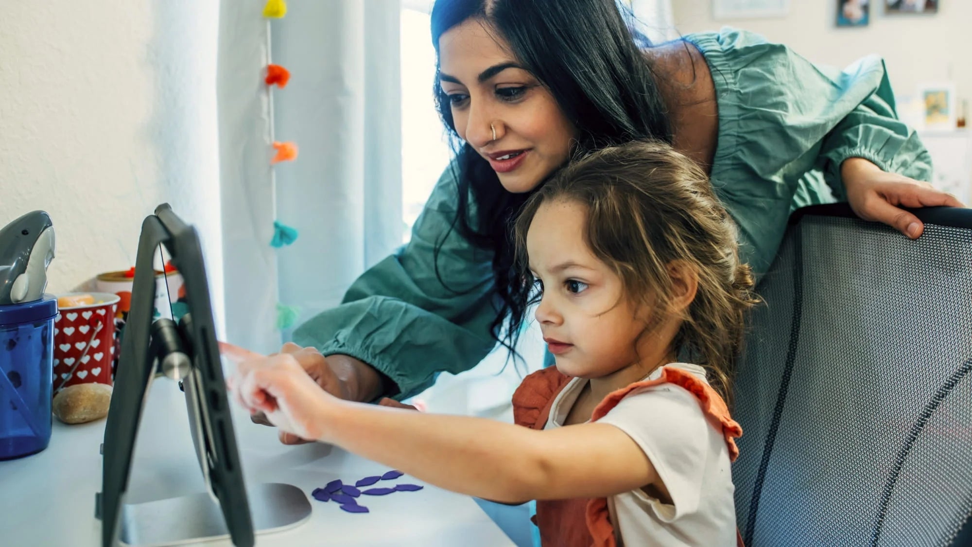 An adult woman helps a young girl using a tablet at a desk. The child points at the screen while the woman leans over, smiling. The scene appears to be indoors with craft supplies nearby.