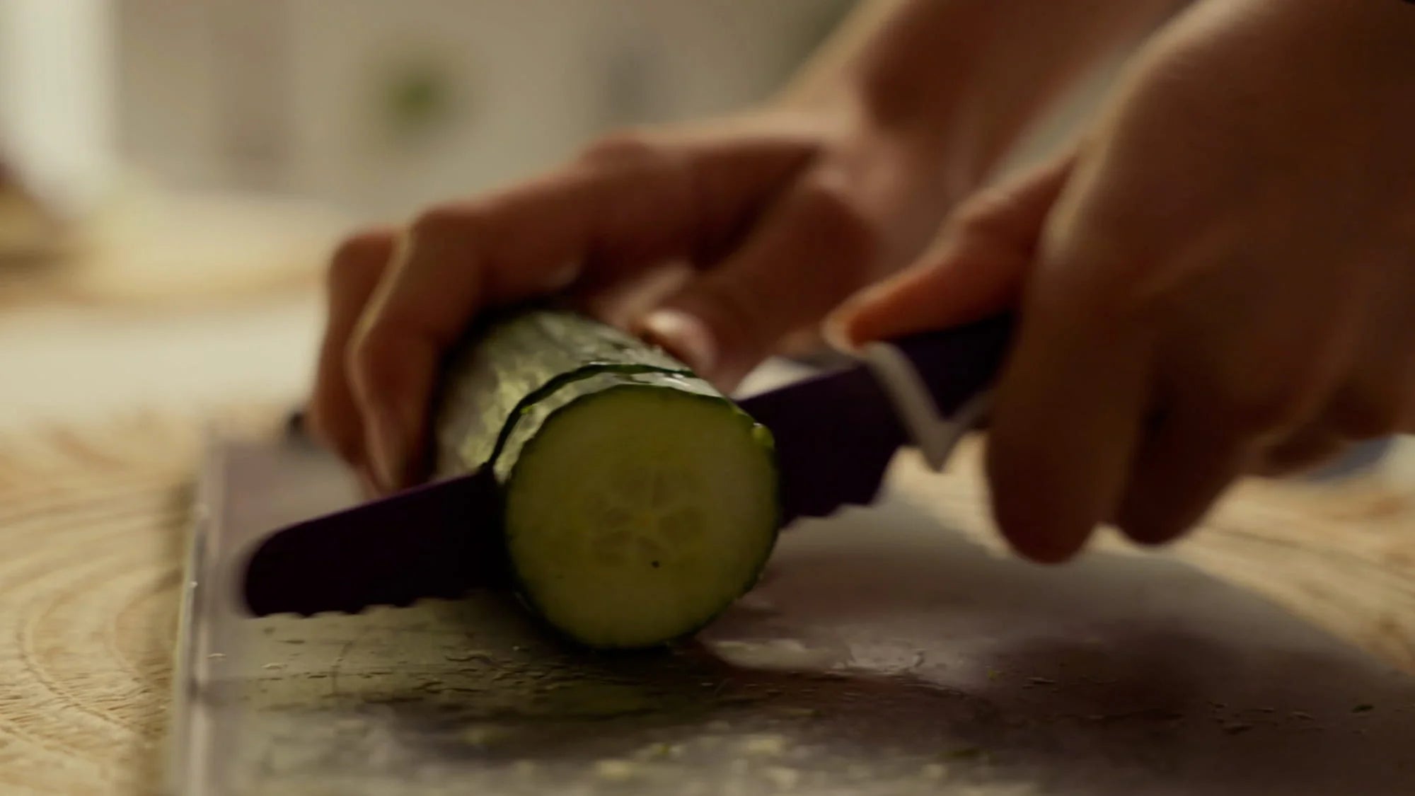 Close-up of a person slicing a cucumber with a purple-handled knife on a cutting board. The hands are holding the cucumber steady while making a cut.