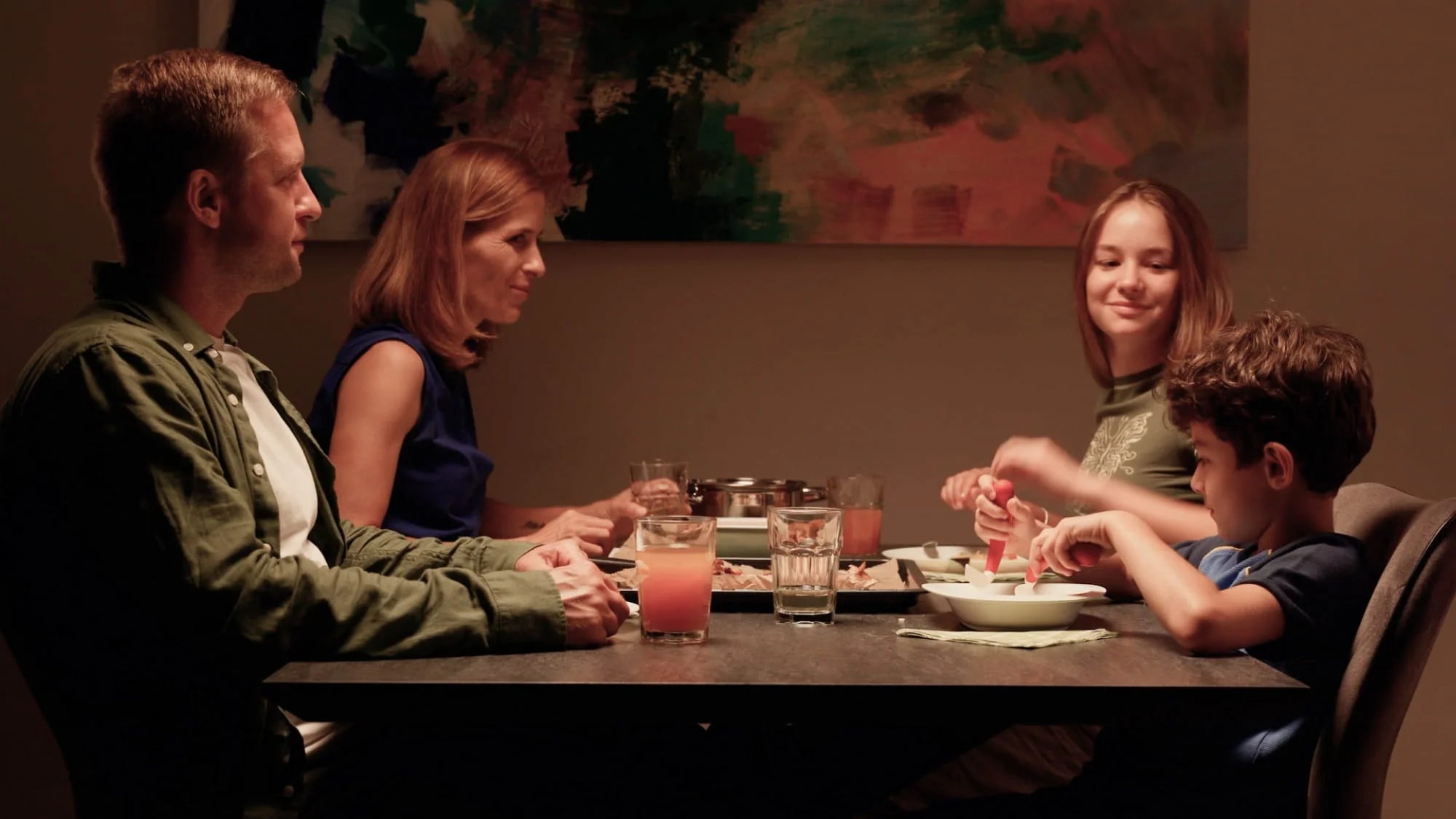 A family of four—two adults and two children—seated around a dining table sharing a meal in a warmly lit room, with drinks and dishes on the table and abstract artwork on the wall behind them.