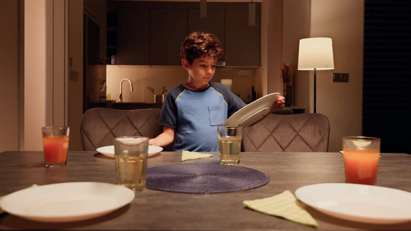 A young boy in a blue shirt sets a plate on a dining table with drinks, plates, and napkins. The room is warmly lit, and the kitchen is visible in the background.