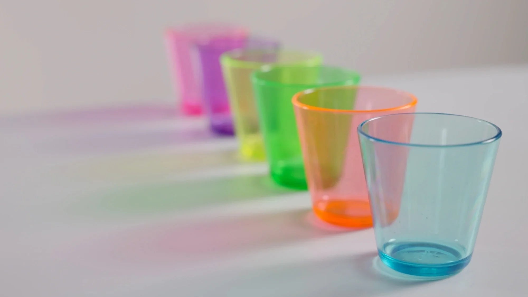 Six translucent plastic cups in blue, orange, green, pink, purple, and red are arranged in a line on a white surface, casting colorful shadows.
