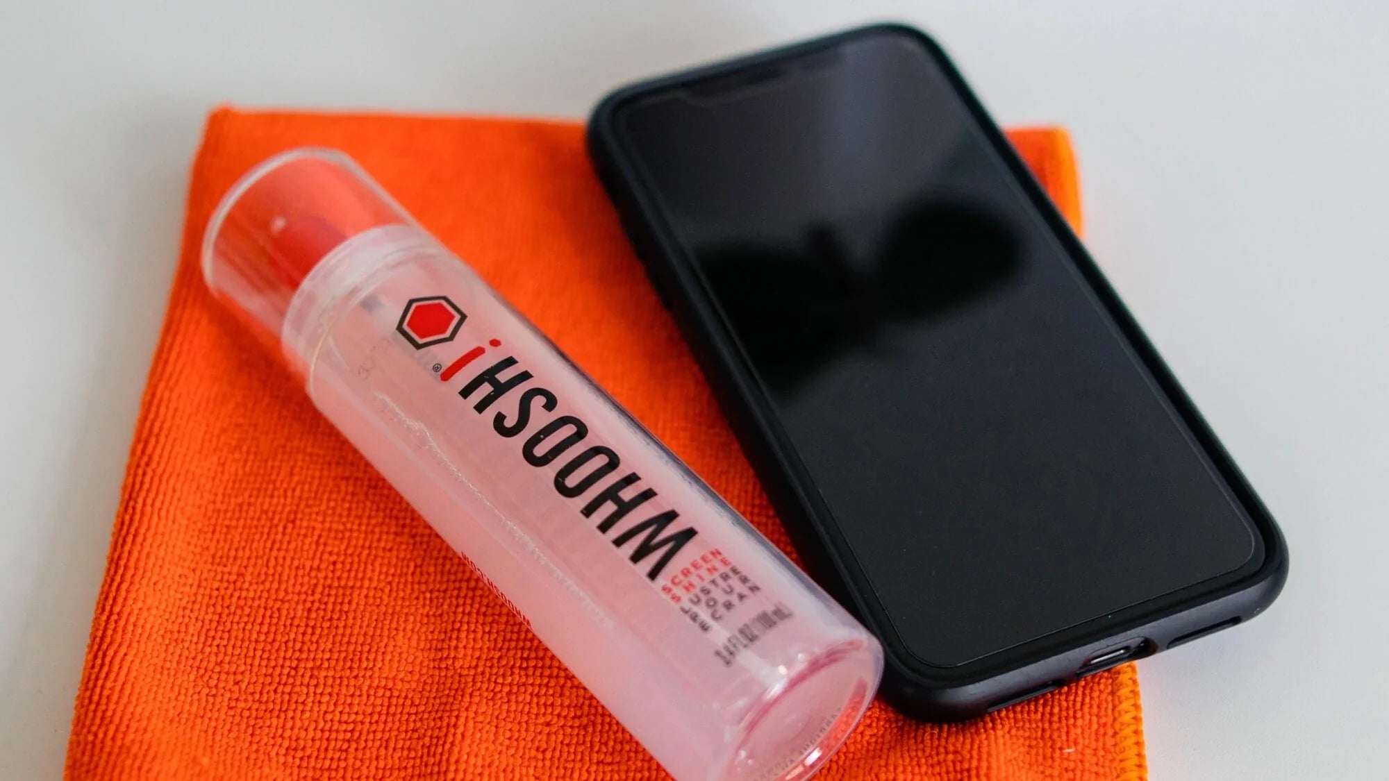 A smartphone, a bottle of WHOOSH! cleaning spray, and an orange microfiber cloth are placed on a white surface.