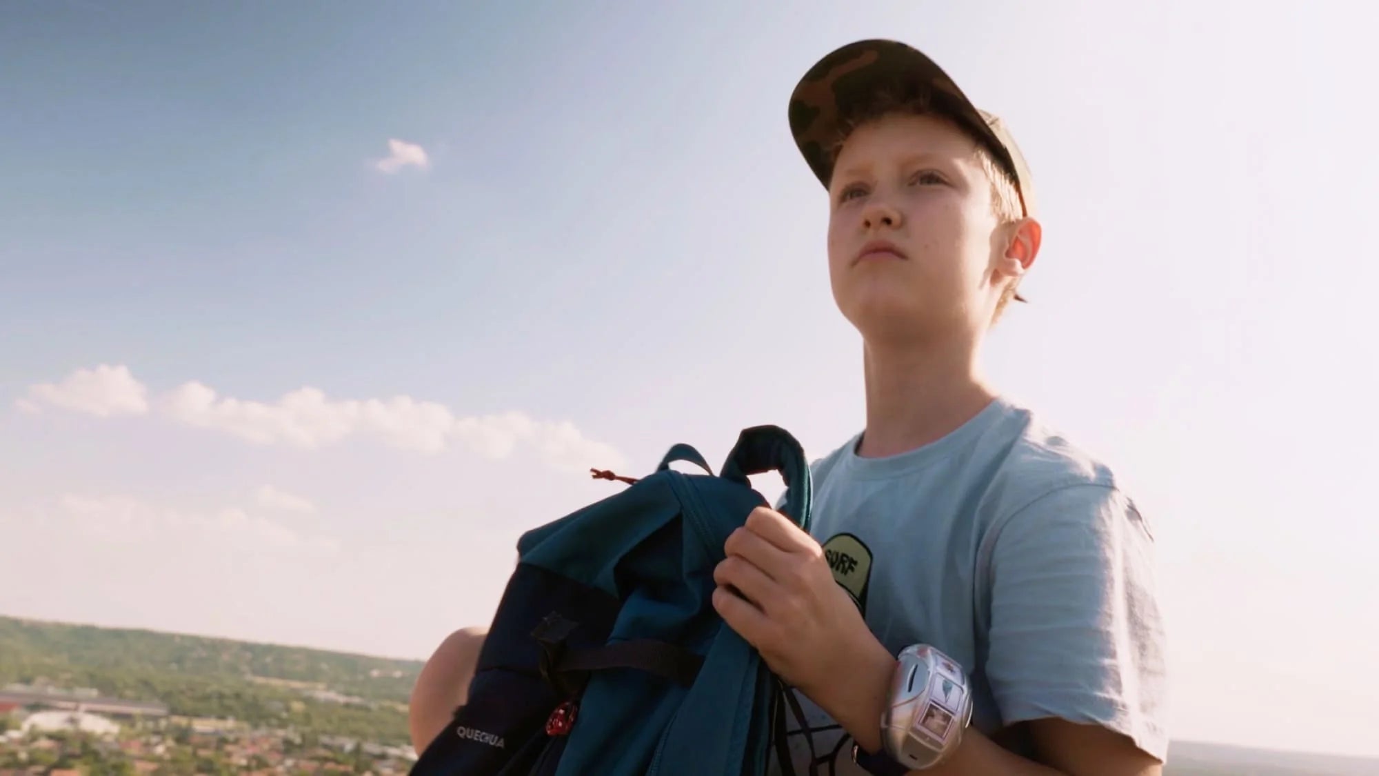 A boy wearing a cap and light blue shirt holds a backpack while looking into the distance, standing outdoors under a bright sky with a landscape in the background.