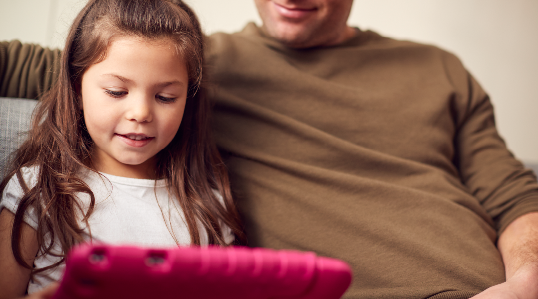 A young girl with long brown hair uses a pink tablet while sitting next to an adult in a brown sweater, both appearing engaged and relaxed on a couch.