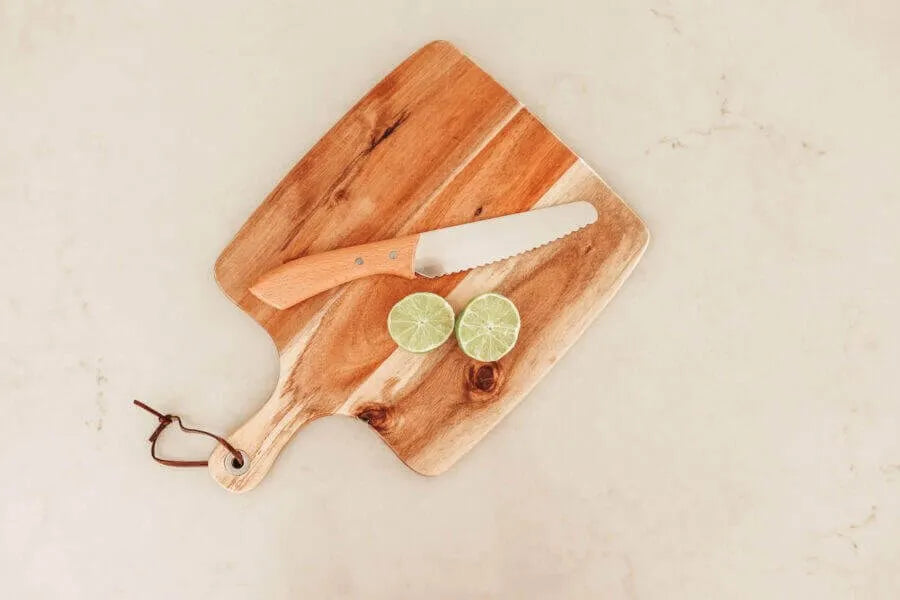 A wooden KandoKutter Adult Safe Knife by KiddiKutter, a cutting board with a brown leather loop, and a halved lime rest on a light marble surface.
