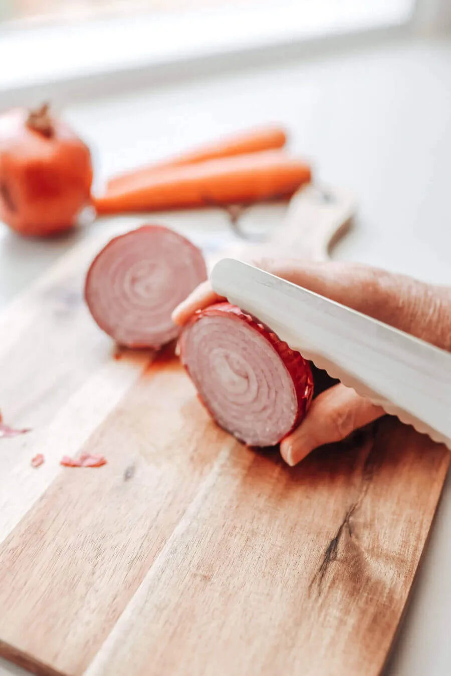 A hand uses the KiddiKutter KandoKutter Adult Safe Knife [Wooden] to slice a red onion on a wooden cutting board, with two carrots and another onion blurred in the background.