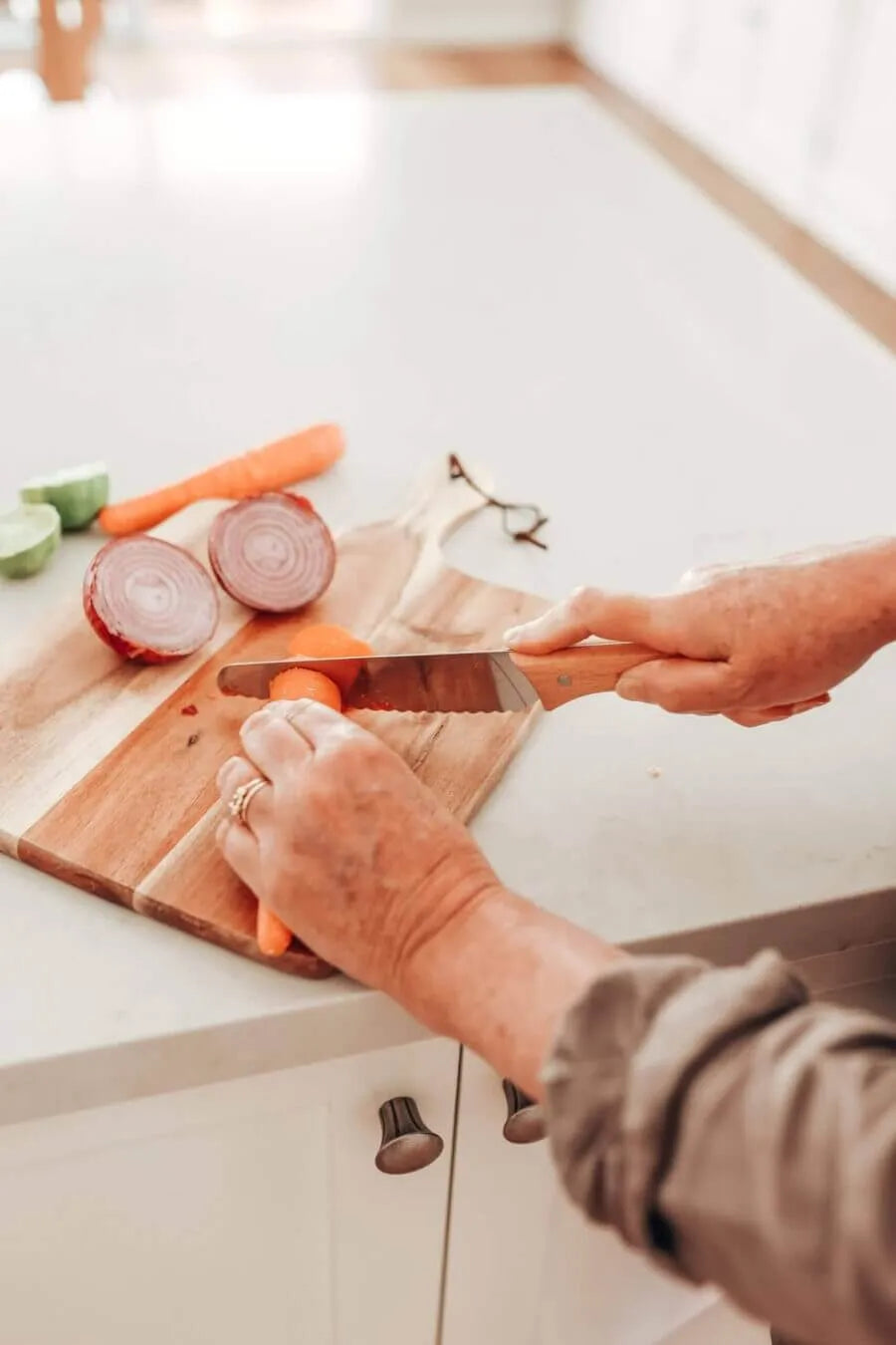 A person uses the KiddiKutter KandoKutter Adult Safe Knife [Wooden] to slice a carrot on a wooden cutting board in a bright kitchen. Sliced red onion, another carrot, and glasses are also on the counter. Only their hands and forearms are visible.