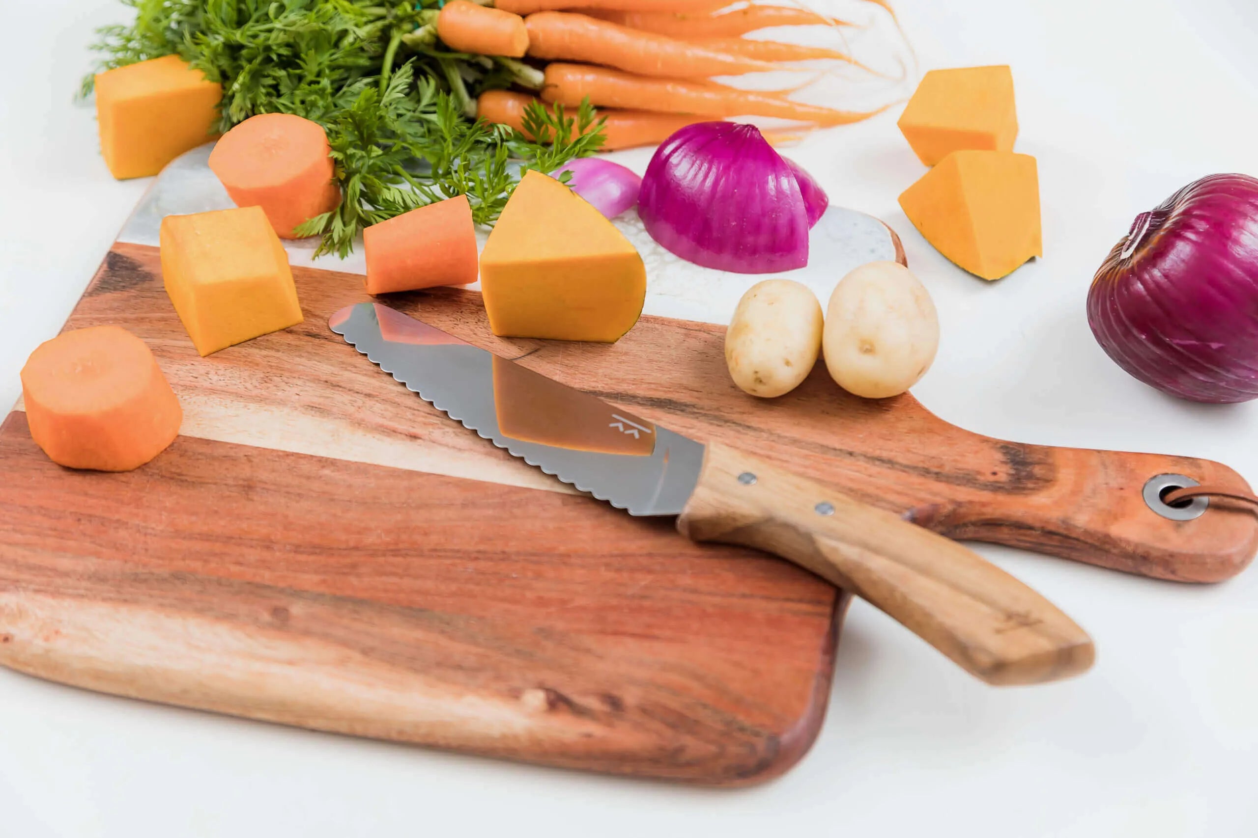 The KandoKutter Adult Safe Knife [Wooden] by KiddiKutter lies on a wooden cutting board surrounded by diced carrots, potatoes, red onion, pumpkin, and whole carrots with greens on a white surface.