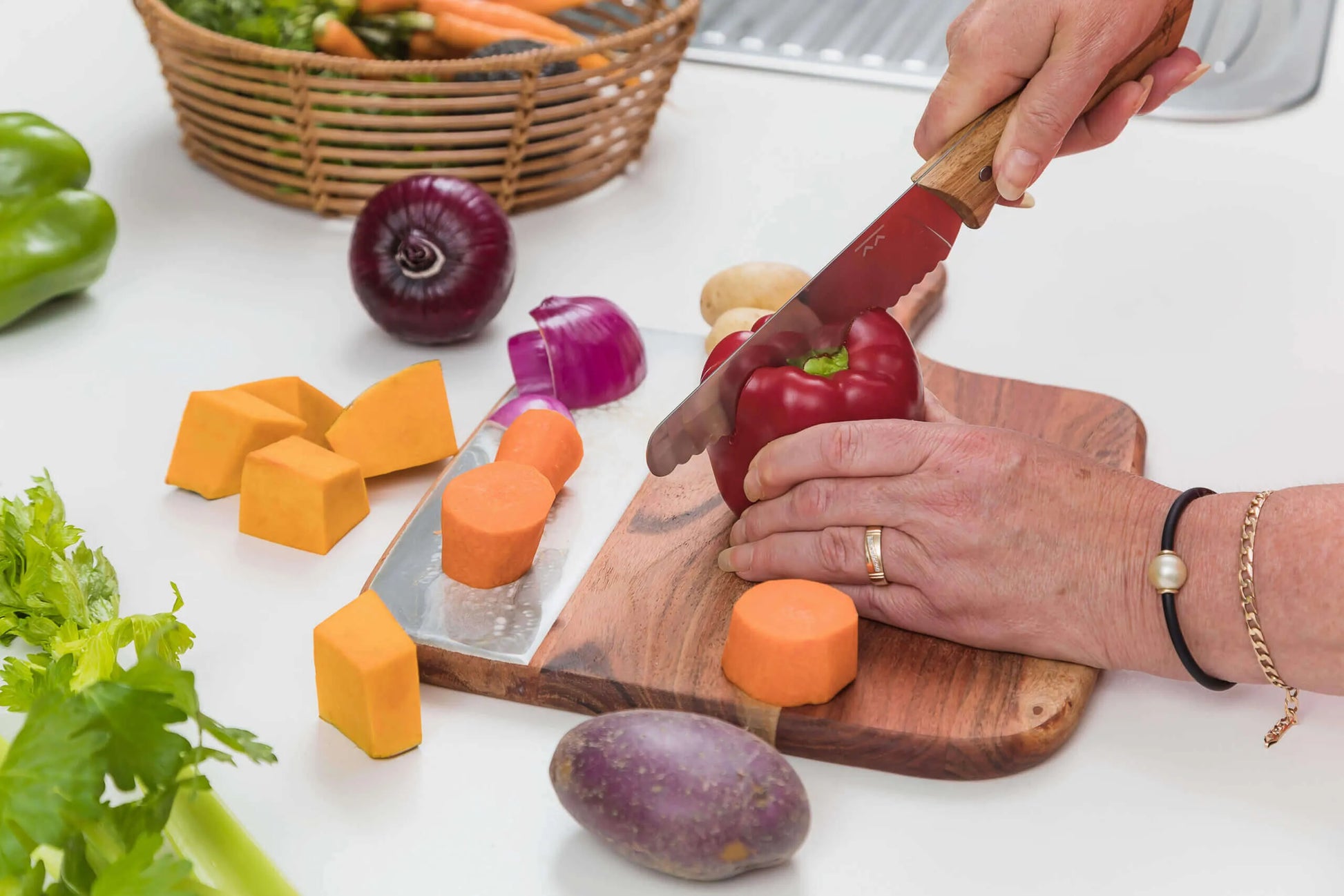 Using the KiddiKutter KandoKutter Adult Safe Knife [Wooden], a person slices a red bell pepper on a wooden cutting board with chopped sweet potatoes, butternut squash, onion, and a basket of vegetables in the background.