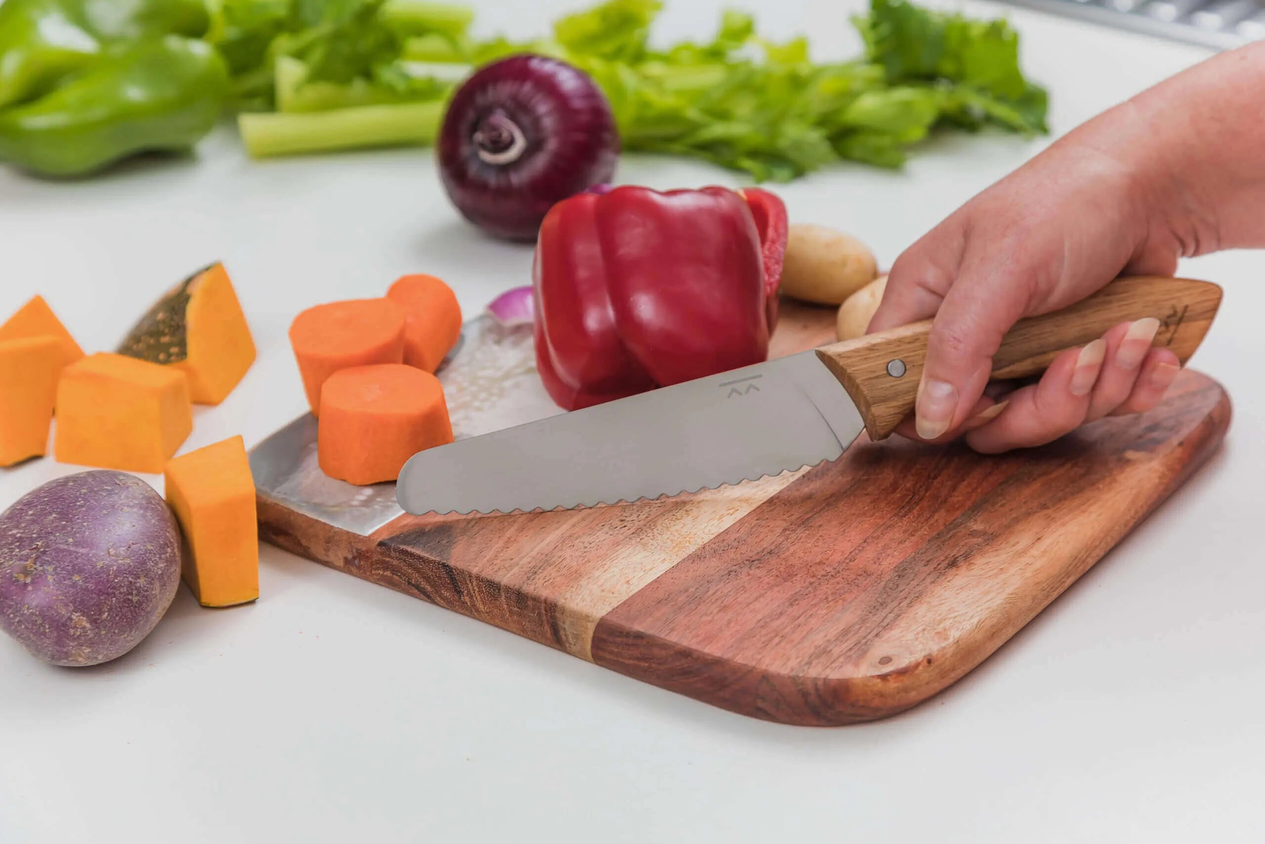 A hand uses the KiddiKutter KandoKutter Adult Safe Knife [Wooden] to slice a red bell pepper on a wooden board surrounded by chopped sweet potato, pumpkin, and fresh vegetables.