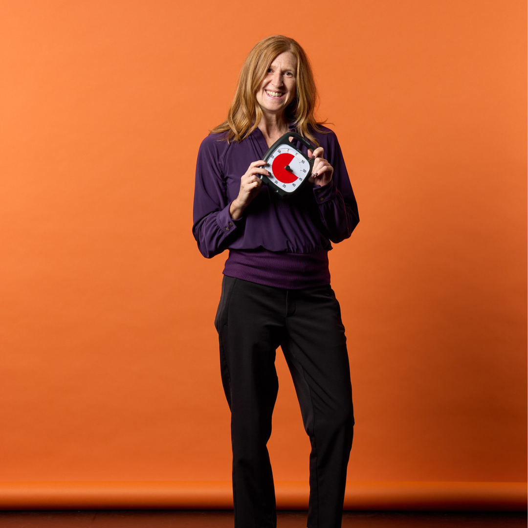 A woman with long, light brown hair in a purple blouse and black pants smiles against an orange background, holding the Time Timer Plus (60 min) by Time Timer—a helpful visual tool for effective time management.