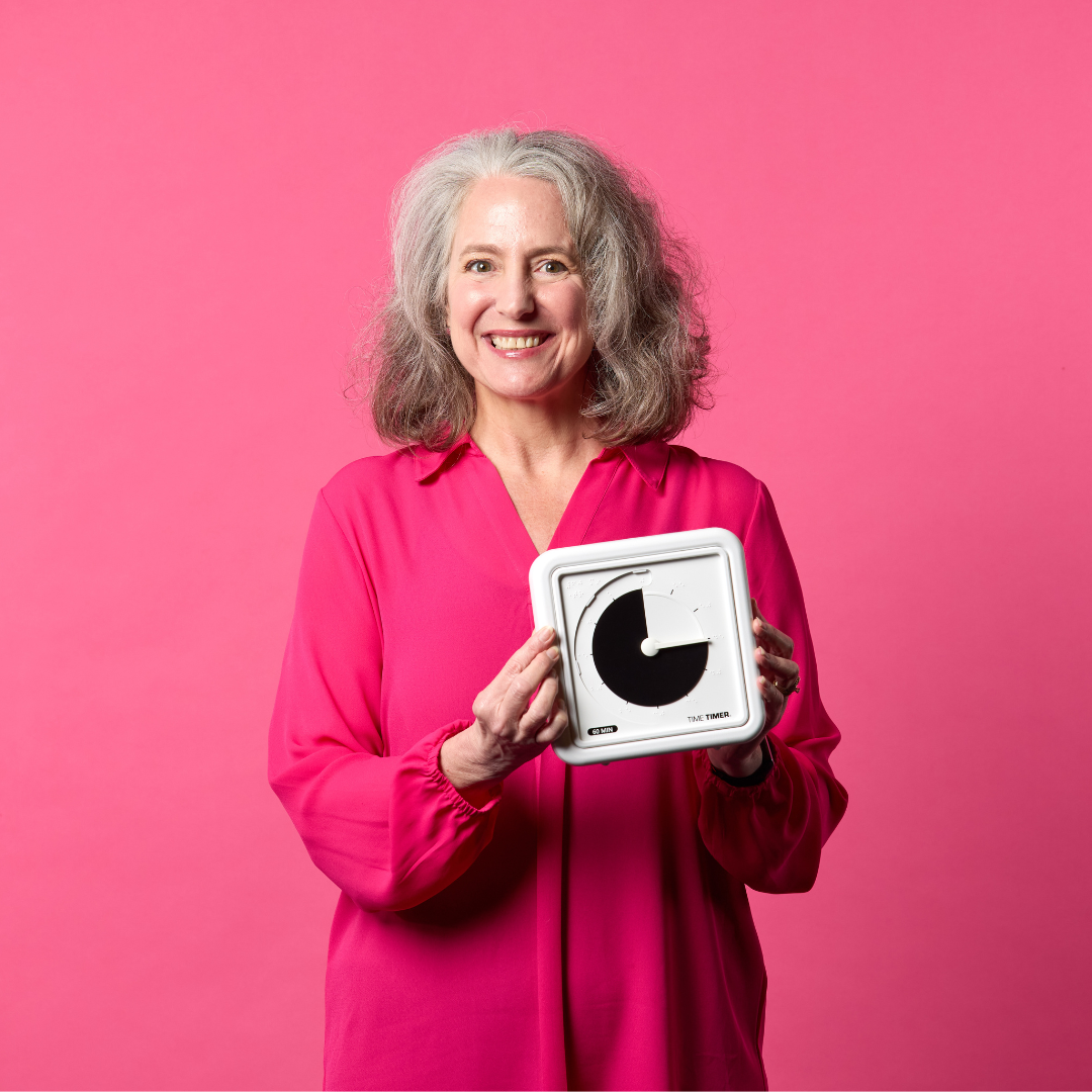 A smiling woman with gray hair in a bright pink blouse stands before a pink background, holding the Time Timer Braille (8 Inch) [Black Disk] by Time Timer, a white tactile timer clock with a minimalist black face.