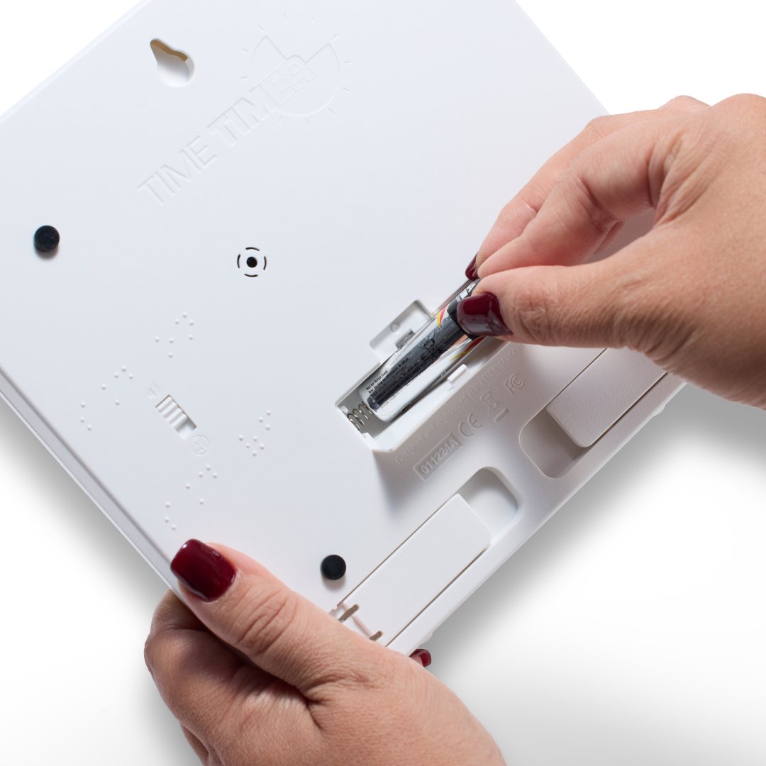 A person with red nail polish inserts an AA battery into the back of a white Time Timer Braille (8 Inch) [Black Disk] device by Time Timer, a tactile timer and popular assistive technology.