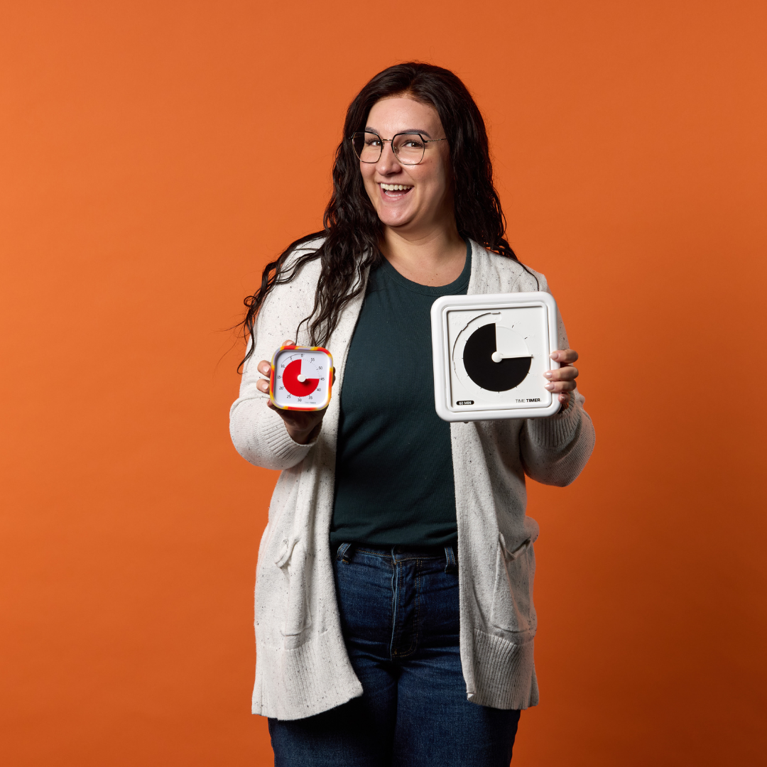 A smiling woman with long dark hair and glasses holds two Time Timer tactile timers, including a Time Timer Braille (8 Inch) [Black Disk] by Time Timer, against an orange background. She wears a gray cardigan, dark green shirt, and blue jeans.