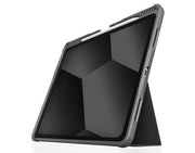 An iPad Air with its screen off is propped in landscape mode using the foldable STM Dux Plus Case by STM, featuring a gray protective design and Apple Pencil storage along the top edge. Geometric shapes are reflected on the display.