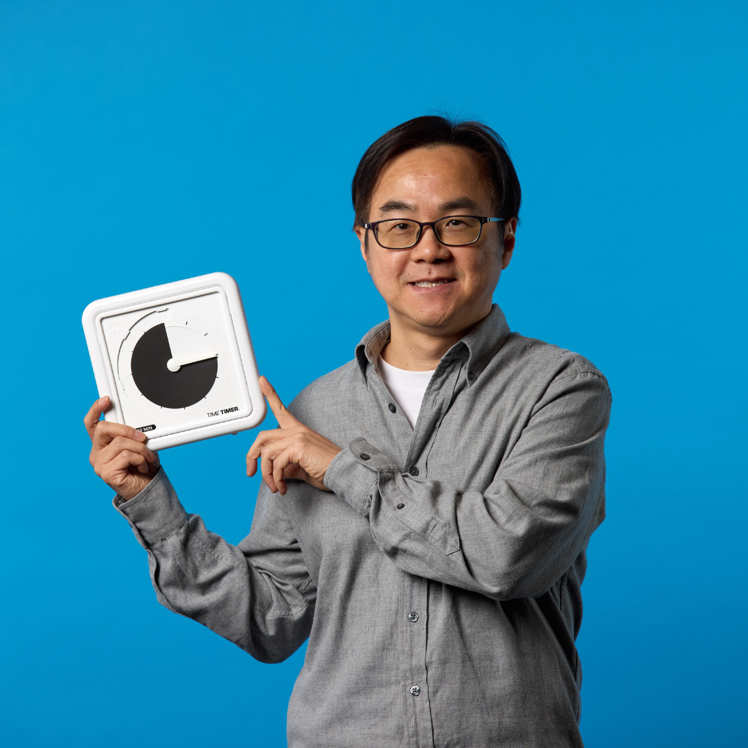 A smiling person in glasses and a gray shirt stands before a blue background, holding and pointing at the Time Timer Braille (8 Inch) [Black Disk] by Time Timer—a tactile assistive timer with a clearly visible large black disk.