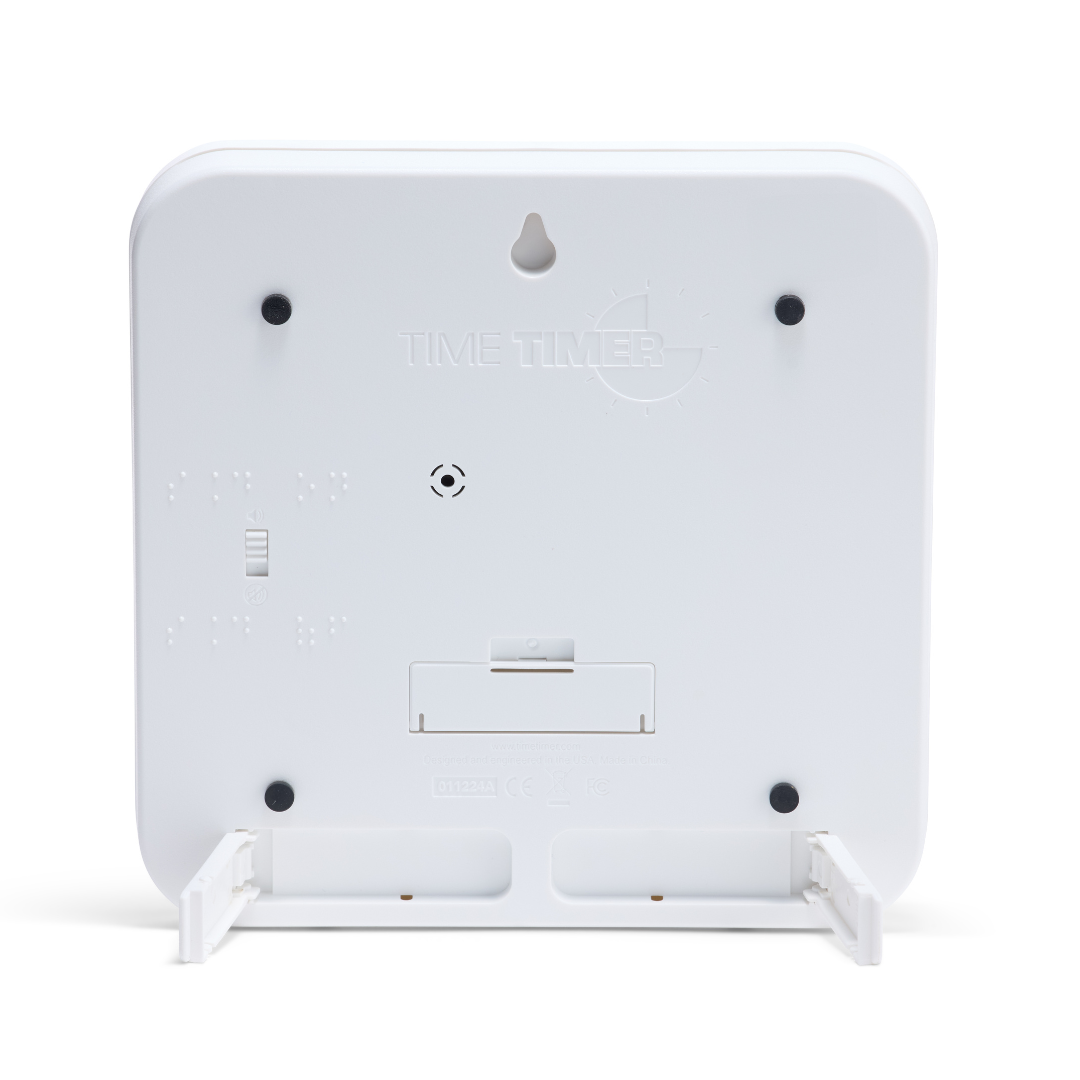 The image shows the back of a white Time Timer Braille (8 Inch) [Red Disk] by Time Timer, featuring braille labels, a battery compartment, small switch, wall-mount hole, and fold-out stands at the base.
