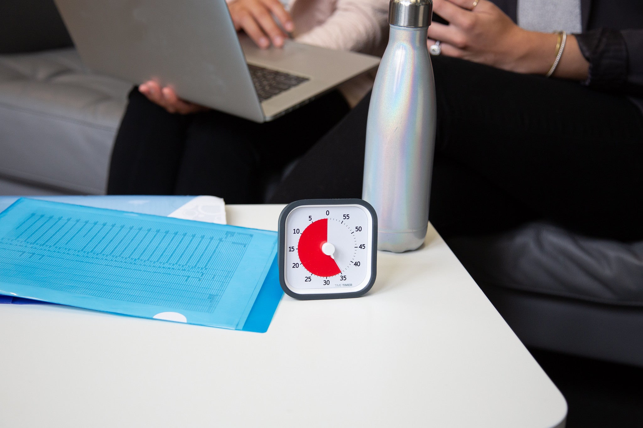 A Time Timer MOD [Sky Blue] (60 min) by Time Timer rests on a white table beside a blue folder and metal water bottle, while two people sit behind on a couch—showcasing this effective time management tool in use.