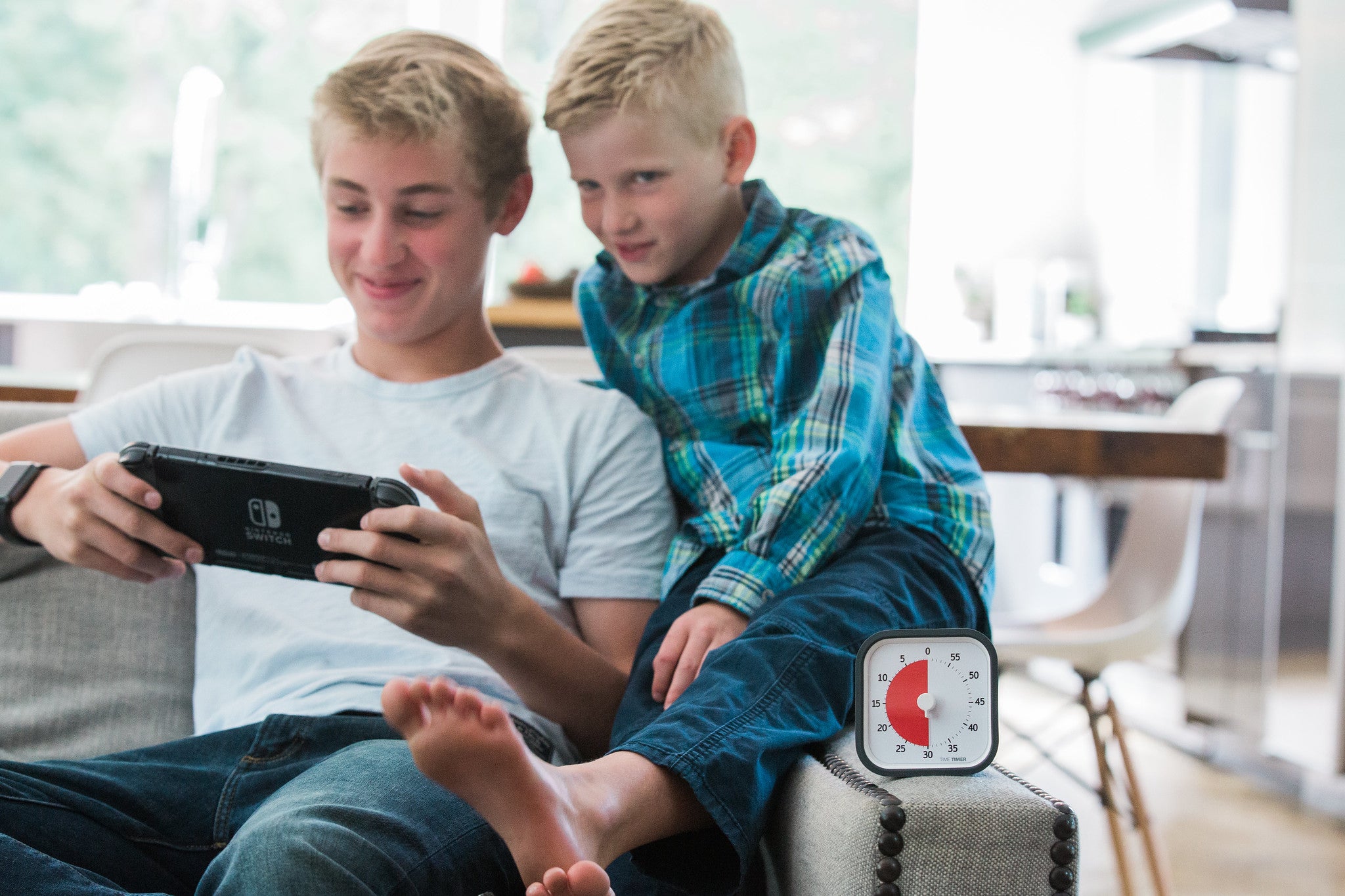 Two boys sit on a couch, smiling as the older one plays a Nintendo Switch. A Time Timer MOD [Charcoal] (60 min) by Time Timer, showing about 40 minutes left, helps them manage playtime. The modern kitchen is bright in the background.