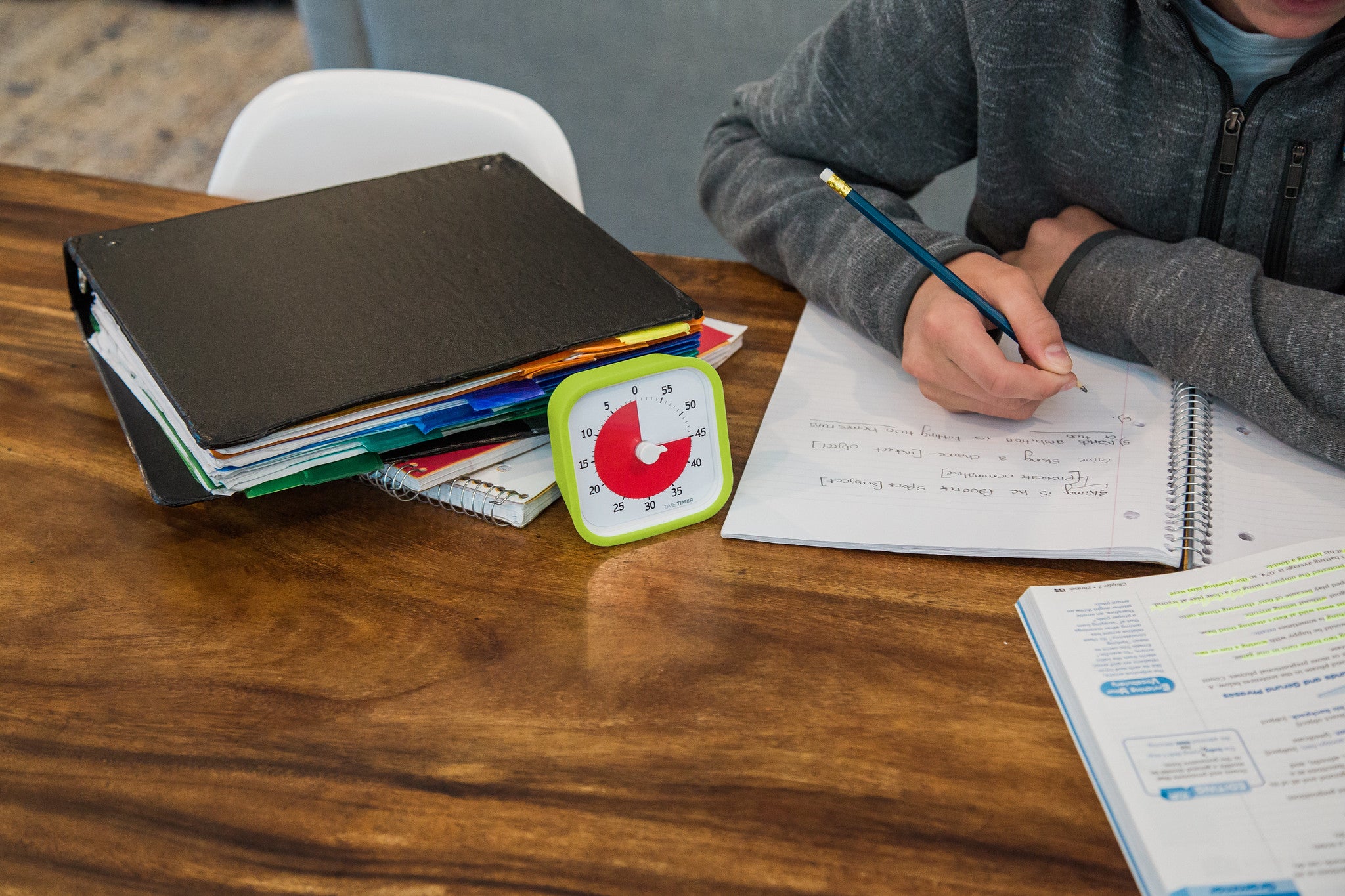 A student writes in a notebook at a wooden table with binders, a book, and a Time Timer MOD [Lime Green] by Time Timer set to 42 minutes—a useful time management tool. Only the student’s arm and part of their torso are visible.