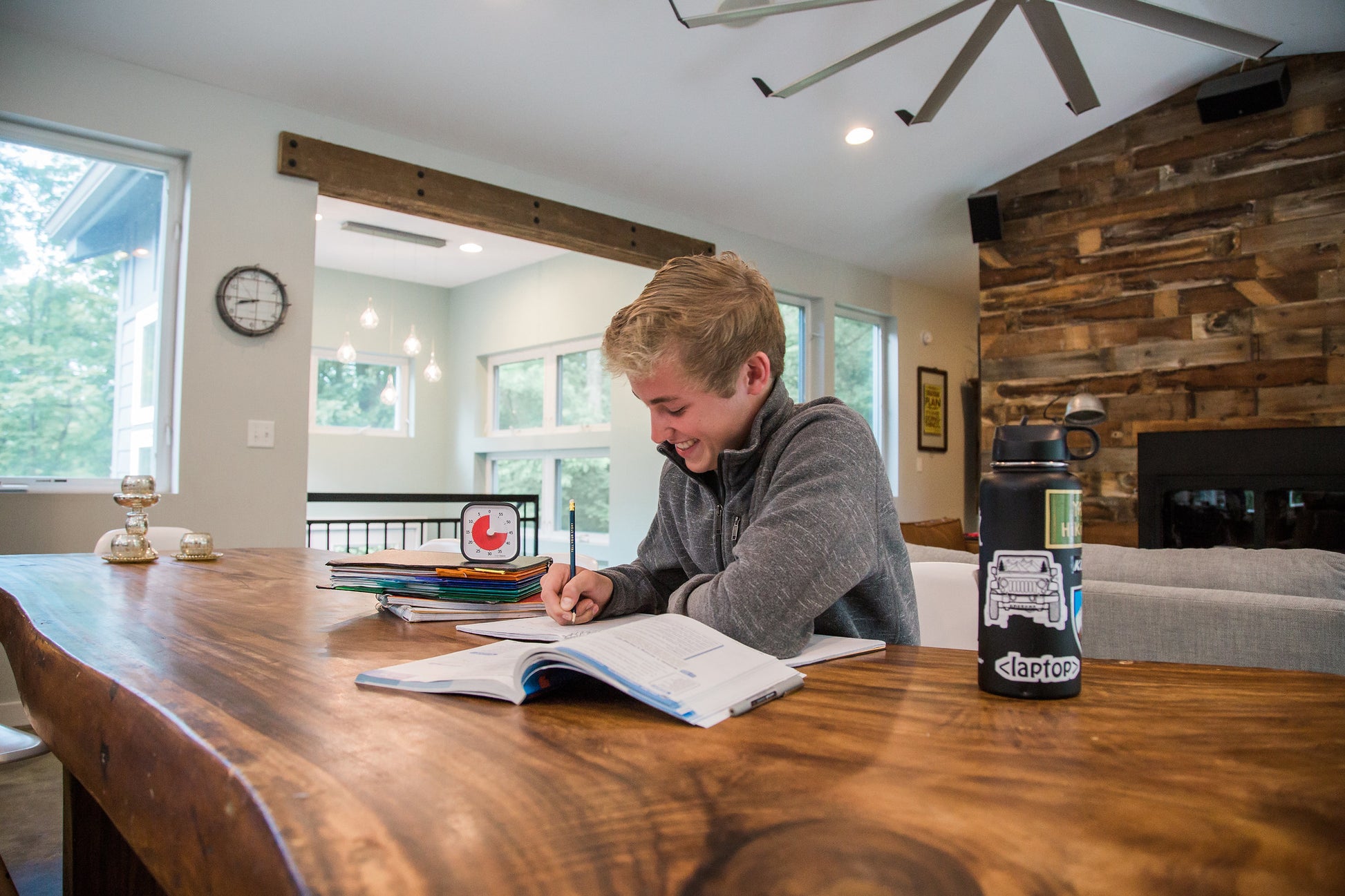 A smiling boy writes in a notebook at a wooden table in a bright, modern room. Nearby are school supplies, papers, folders, a water bottle, and the Time Timer MOD [Charcoal] 60-min visual timer by Time Timer to help manage time.