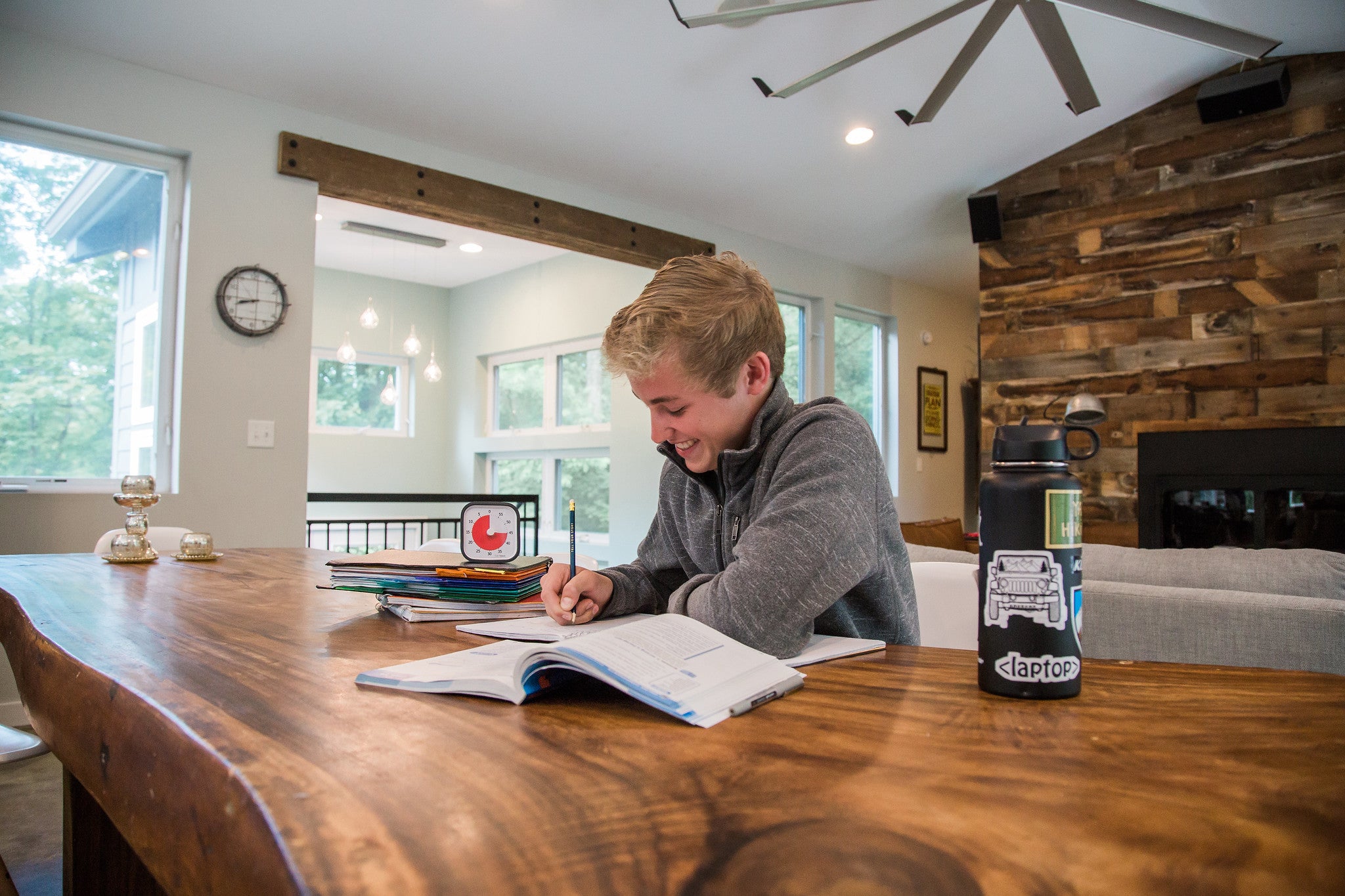 A smiling boy writes in a notebook at a wooden table in a bright, modern room. Nearby are school supplies, papers, folders, a water bottle, and the Time Timer MOD [Charcoal] 60-min visual timer by Time Timer to help manage time.