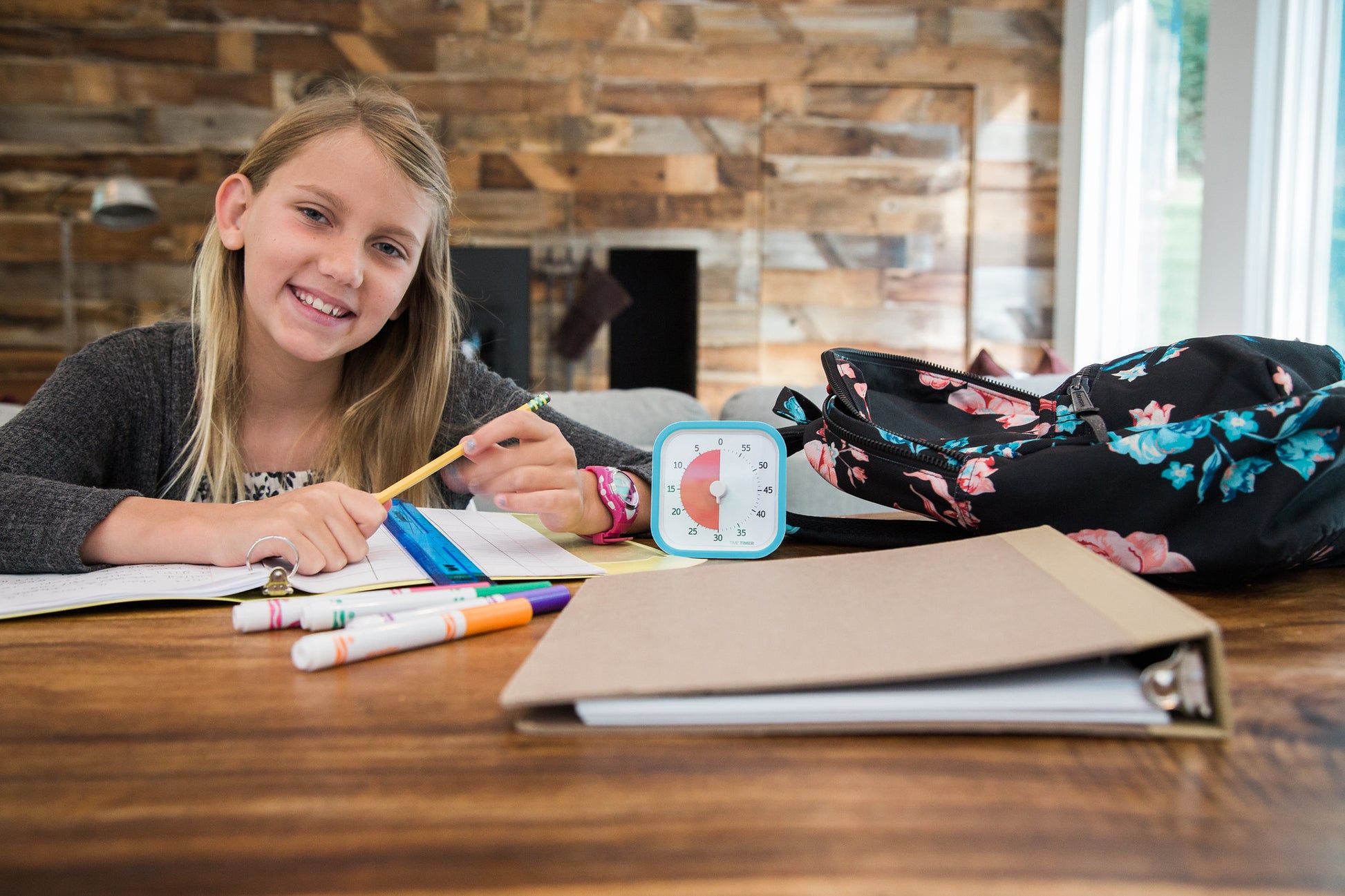 A smiling girl does homework at a table with markers, papers, a binder, a floral backpack, and the Time Timer MOD [Sky Blue] (60 min) by Time Timer to help manage her time. A wooden wall is visible in the background.