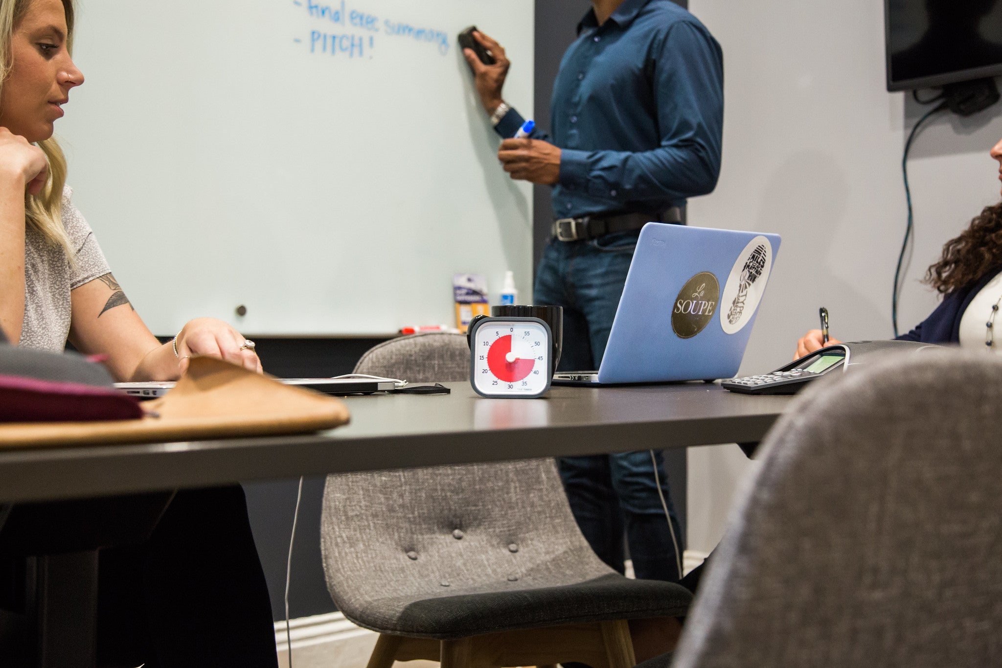 Three people meet in a room; one writes notes on a whiteboard while the others use laptops. A Time Timer MOD [Charcoal] (60 min) by Time Timer sits on the table, helping manage time effectively during their session.