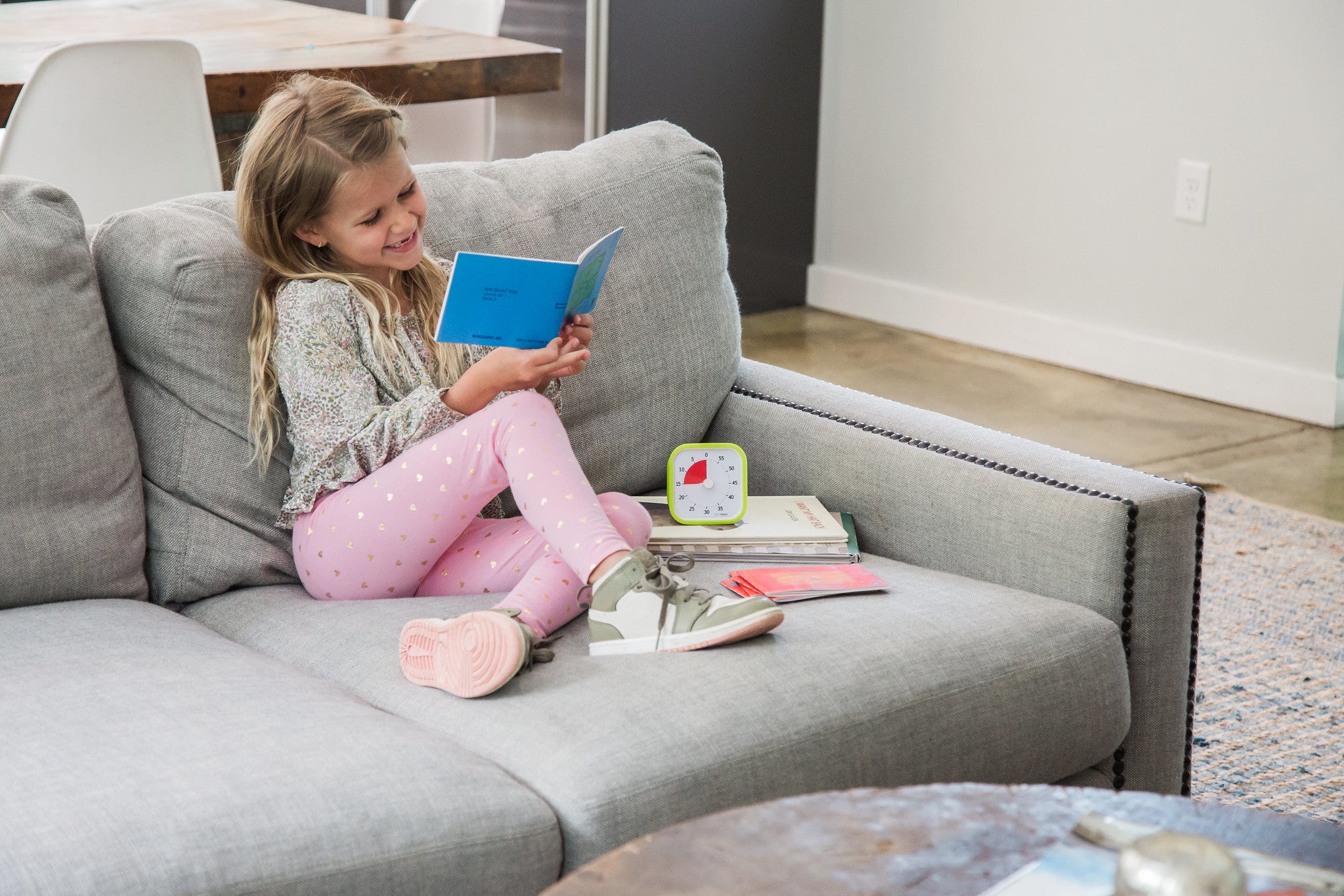 A young girl sits on a gray sofa, smiling as she reads a blue book. She wears pink pants with gold polka dots and sneakers. Beside her are books and the Time Timer MOD [Lime Green] by Time Timer, supporting her time management skills.
