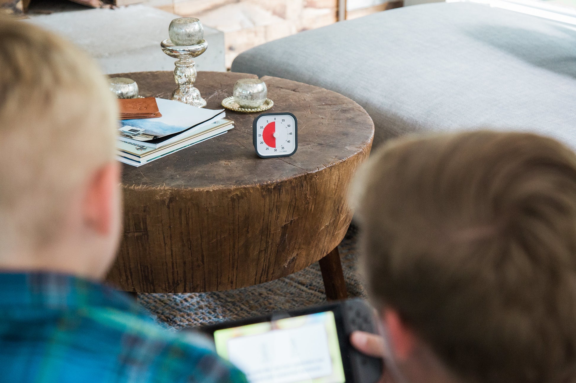 Two children sit near a round wooden coffee table topped with a Time Timer MOD [Charcoal] (60 min) by Time Timer, books, and candle holders. The timer's red disk shows time remaining. They focus on a handheld device; a gray couch is in the background.