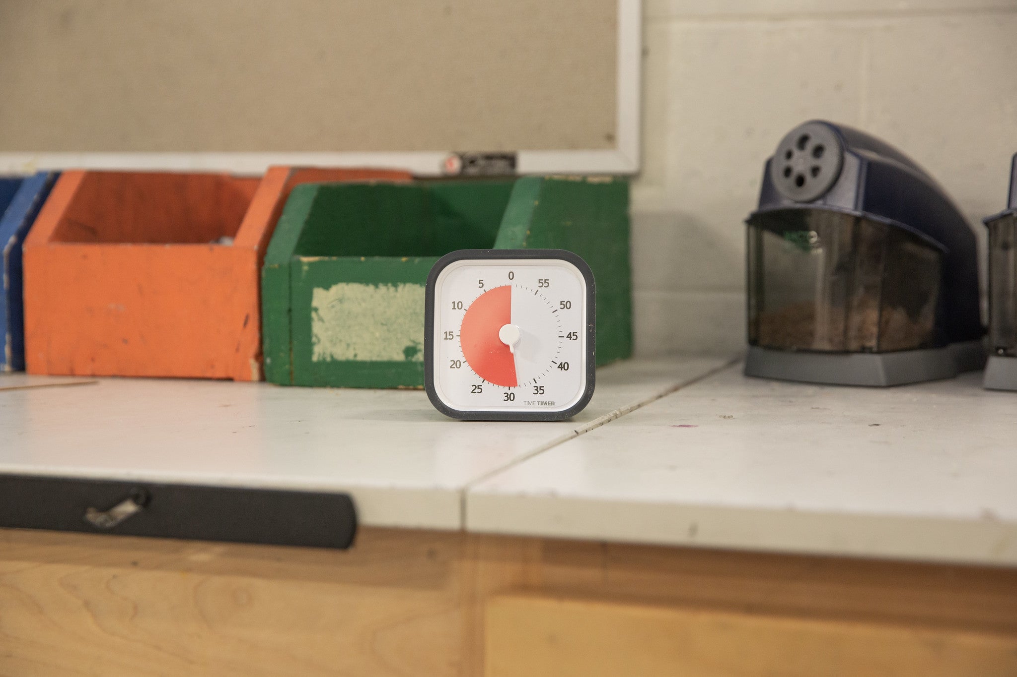 A Time Timer MOD [Charcoal] by Time Timer, set to 30 minutes, sits on a white countertop with orange, green, and blue wooden boxes and a food-filled plastic container against a white wall.