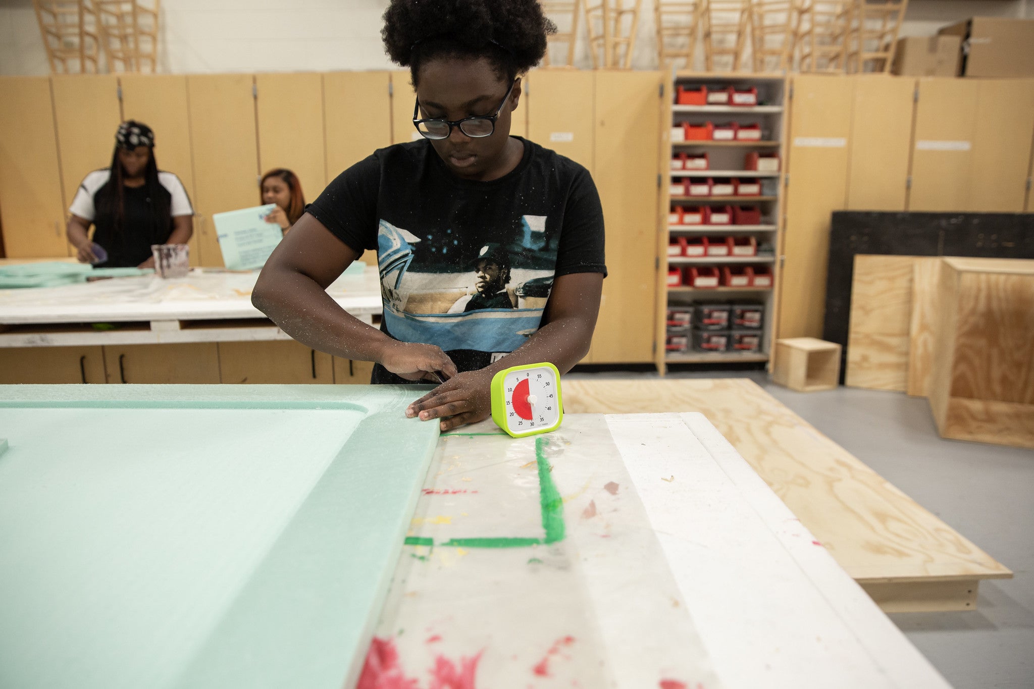 A person wearing glasses works with foam at a table in a workshop. A Time Timer MOD [Lime Green] (60 min) from Time Timer sits nearby, aiding time management. Others and shelves with bins are visible in the background.