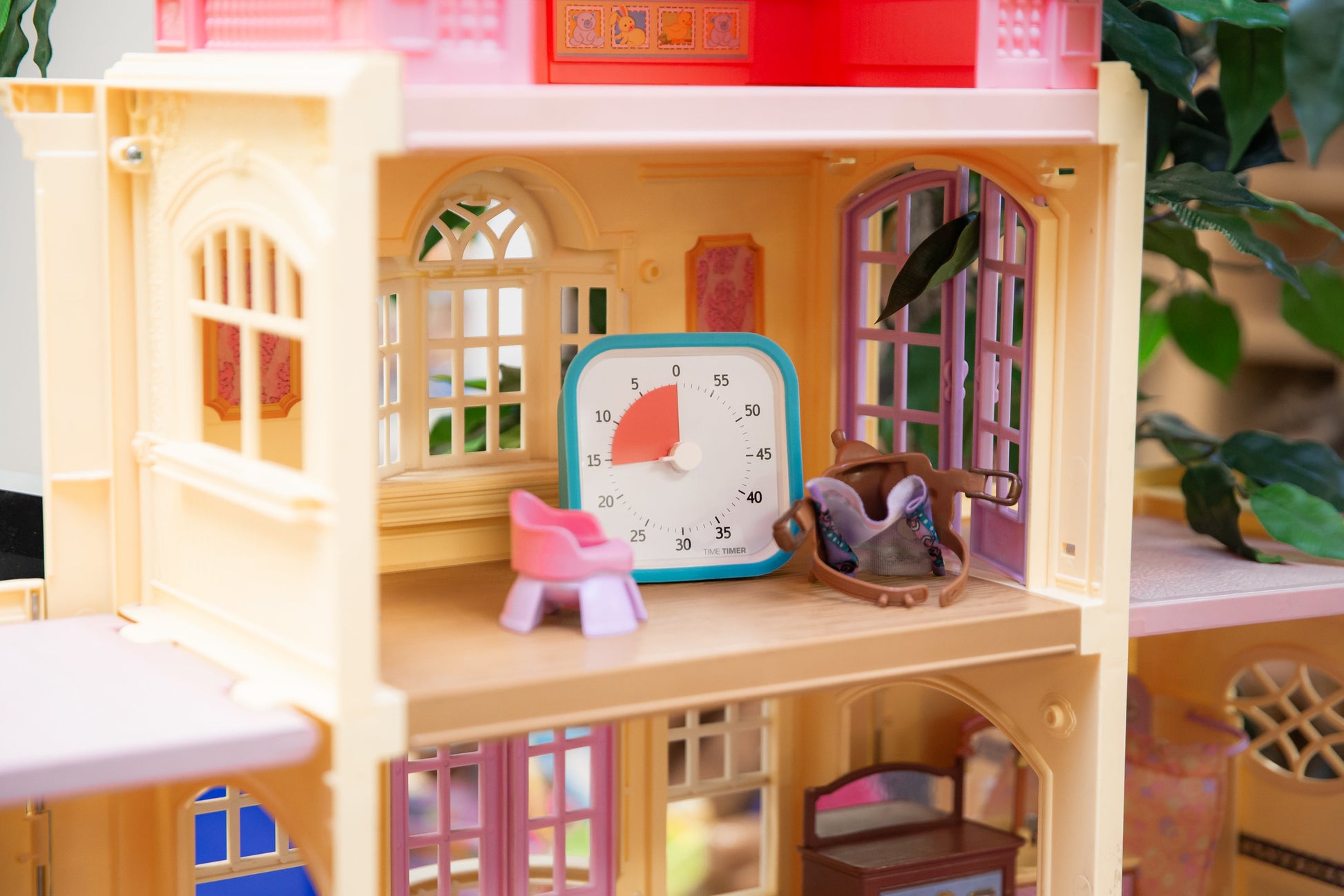 A close-up of a dollhouse room displays a toy pink chair, rocking horse, and a Time Timer MOD [Sky Blue] by Time Timer set to 20 minutes—an ideal playful time management tool. The brightly lit scene is decorated with plants.