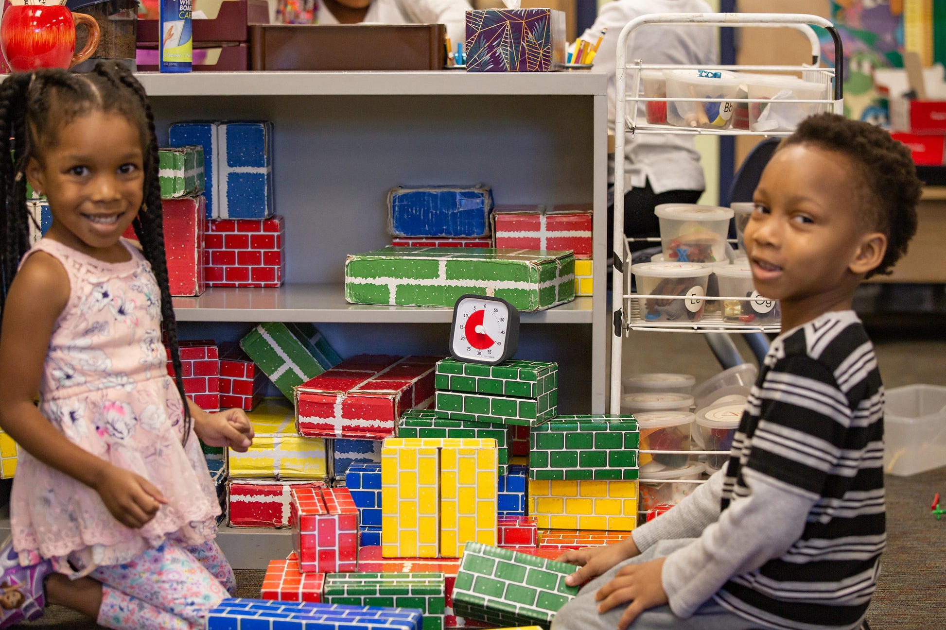 Two young children smile and play with colorful blocks in a classroom. A Time Timer MOD [Charcoal] (60 min) by Time Timer sits nearby, helping teach time management among shelves filled with art supplies in the background.