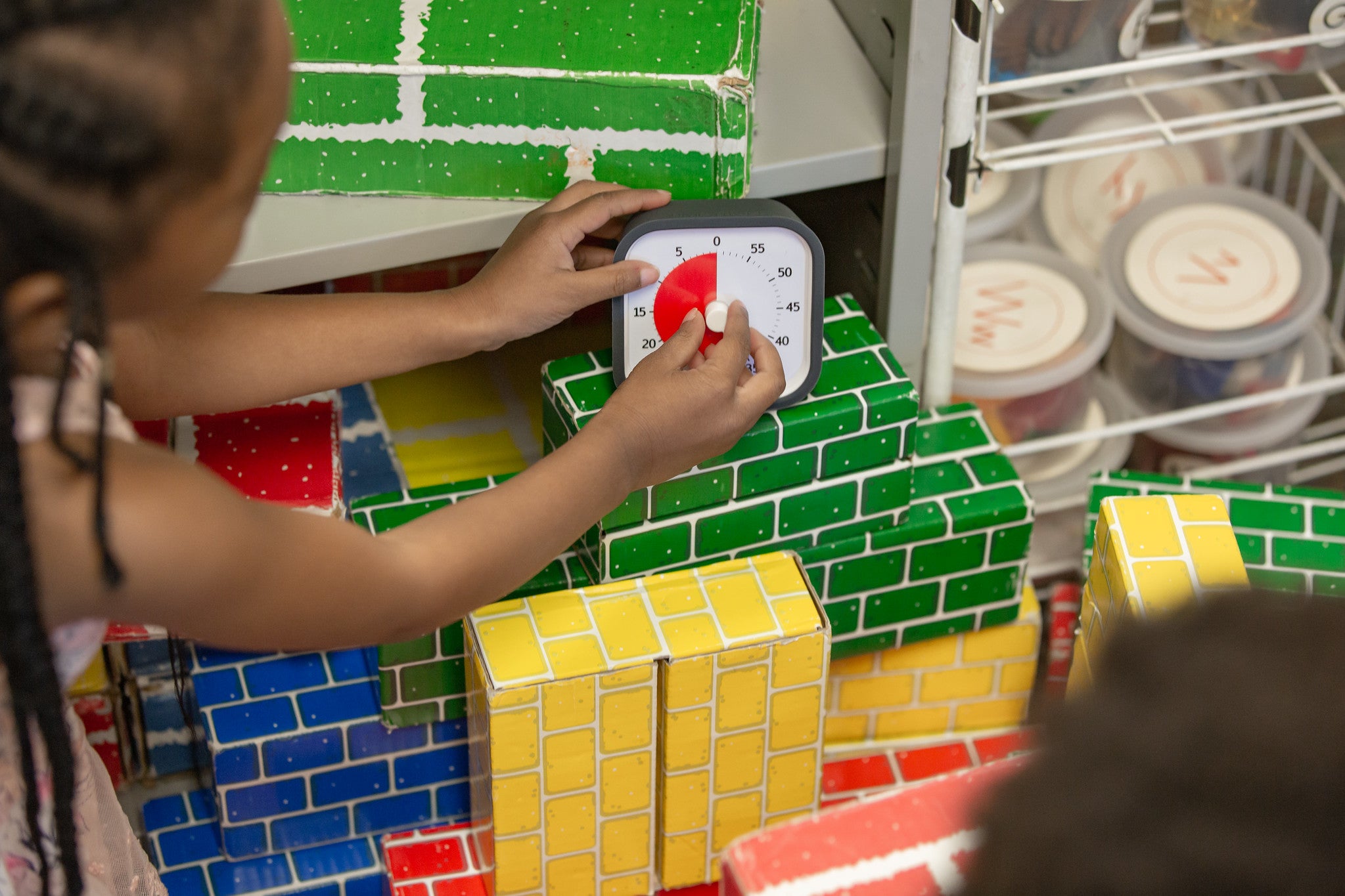 A child places the Time Timer MOD [Charcoal] (60 min) by Time Timer on colorful, brick-patterned blocks in a play area, with shelves and storage containers visible in the background.