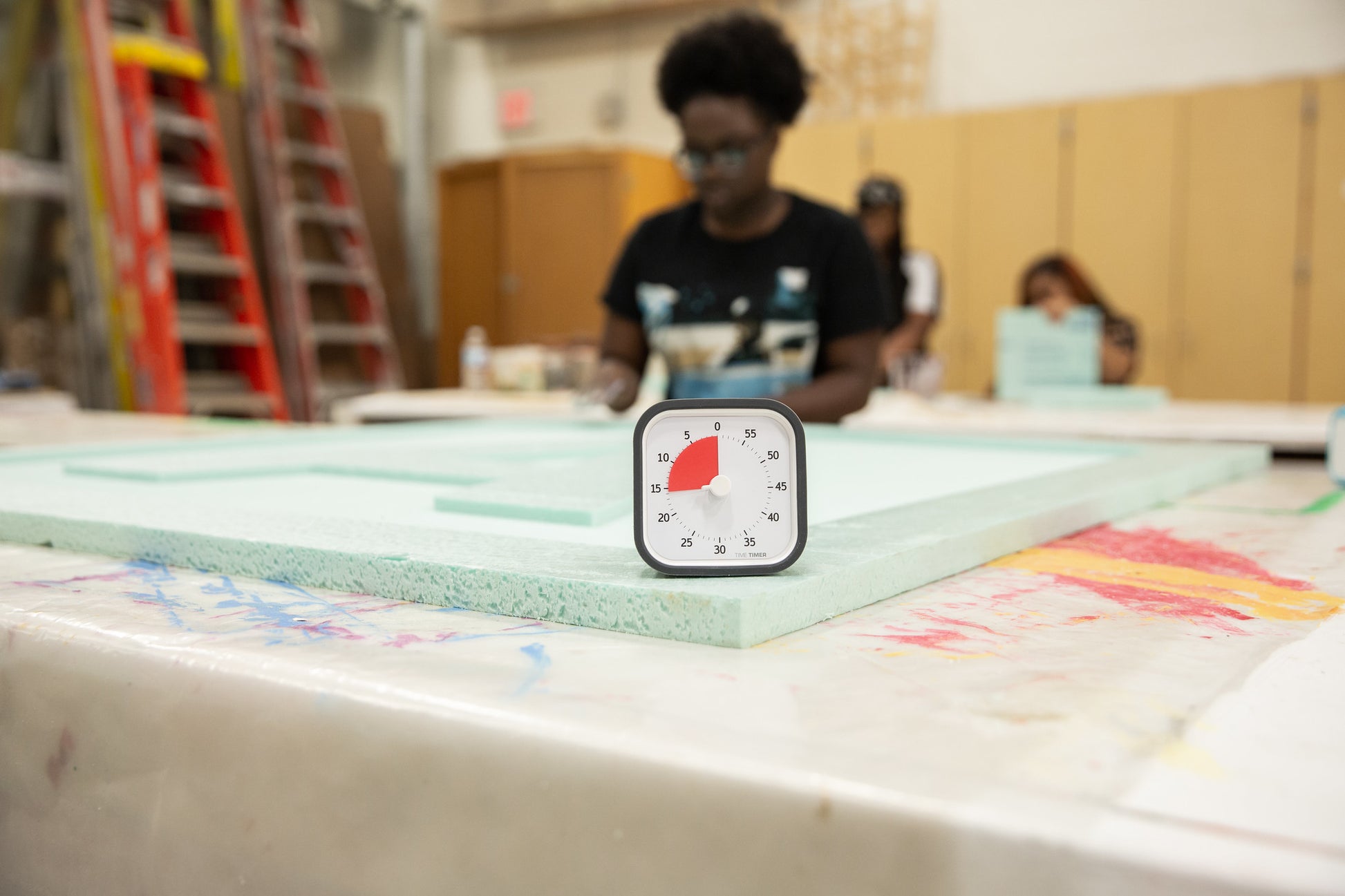 A Time Timer MOD [Charcoal] (60 min) by Time Timer sits on a paint-splattered table set to about 15 minutes, while someone works on a large foam board in the workshop background.