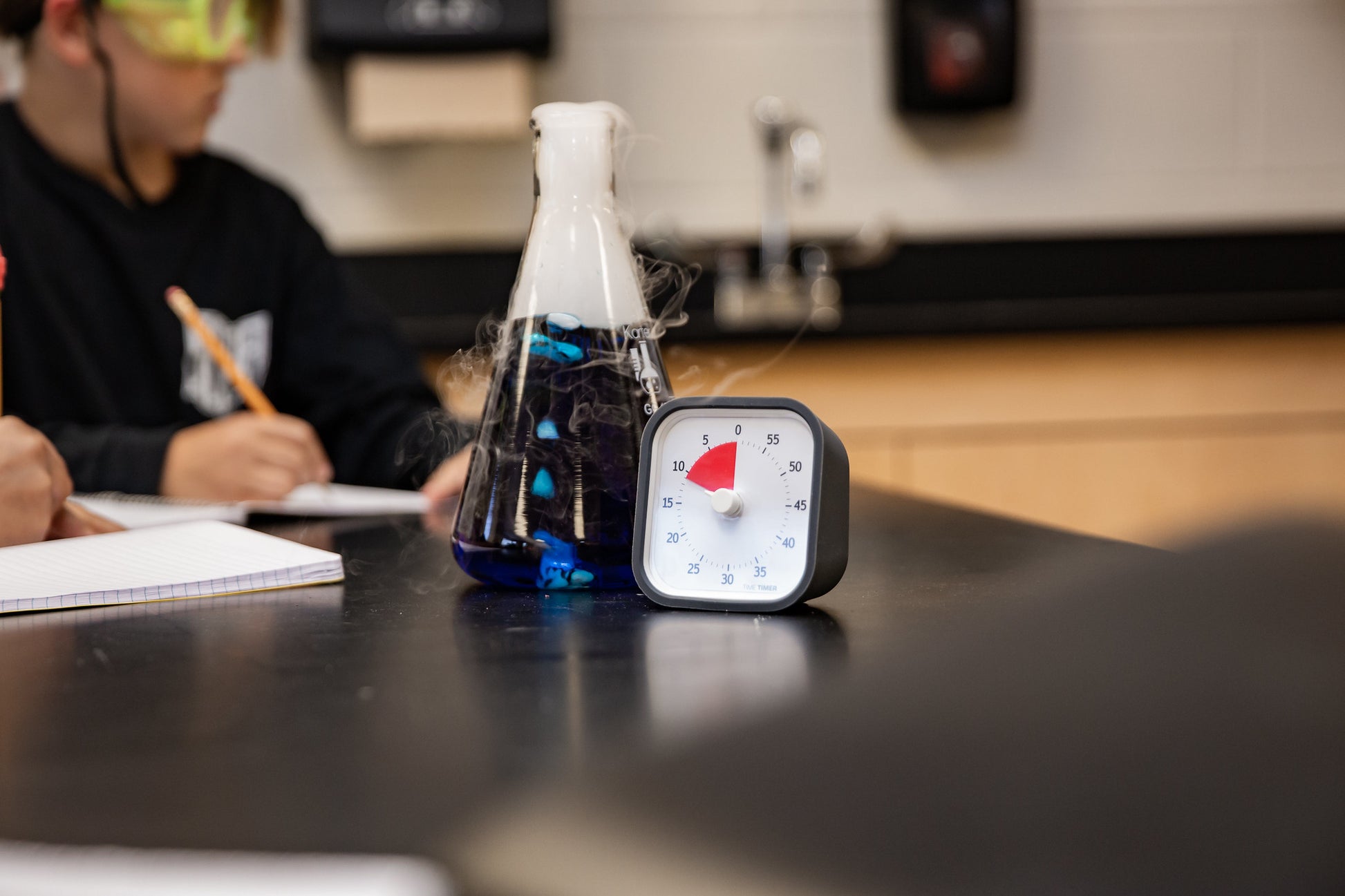 A glass flask with blue liquid and vapor sits beside the Time Timer MOD [Charcoal] (60 min) by Time Timer on a classroom table, while a student writes in a notebook in the background.