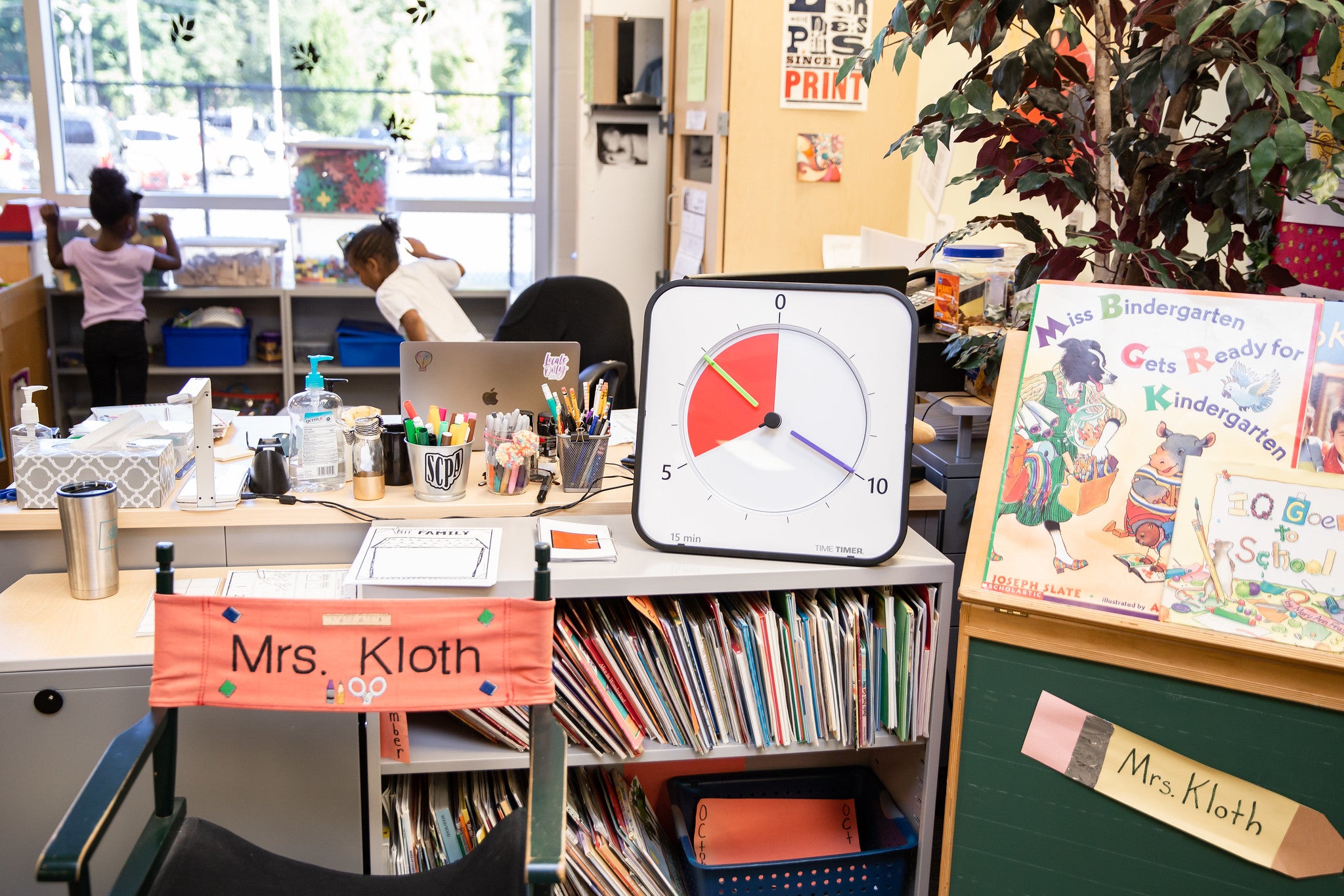 A kindergarten classroom with a teacher’s chair labeled Mrs. Kloth, a desk with the Time Timer MAX and Time Timer MAX Extra Set Markers (3 Colours), bookshelves, two kids by the window, and a display reading "Mrs. Bindergarten Gets Ready for Kindergarten.