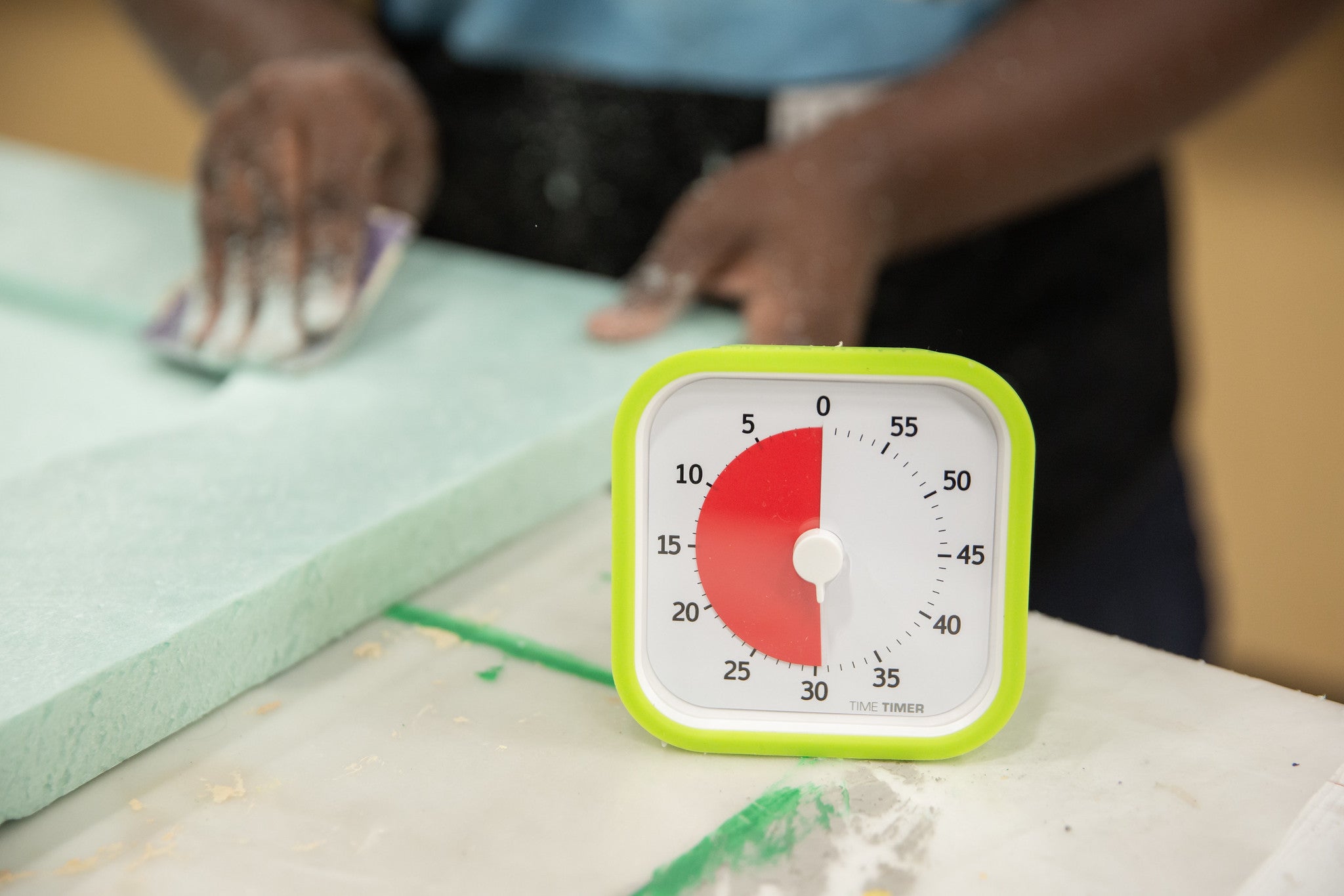 A close-up of the Time Timer MOD [Lime Green] (60 min) by Time Timer, set to 35 minutes with a red time display. In the background, a person sands light green foam, using this timer for efficient work intervals.