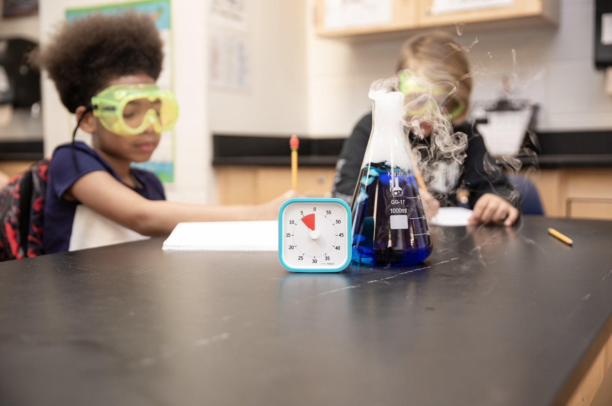 Two children in safety goggles work at a science lab table. On the table is a Time Timer MOD [Sky Blue] (60 min) by Time Timer in a silicone case and a smoking beaker with blue liquid. The kids are taking notes.