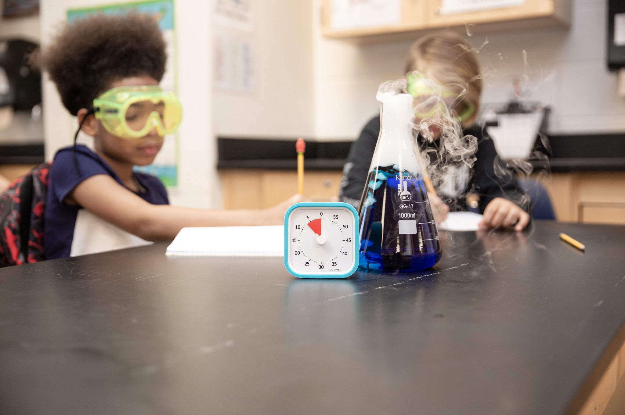 Two children in safety goggles work at a science lab table. On the table is a Time Timer MOD [Sky Blue] (60 min) by Time Timer in a silicone case and a smoking beaker with blue liquid. The kids are taking notes.